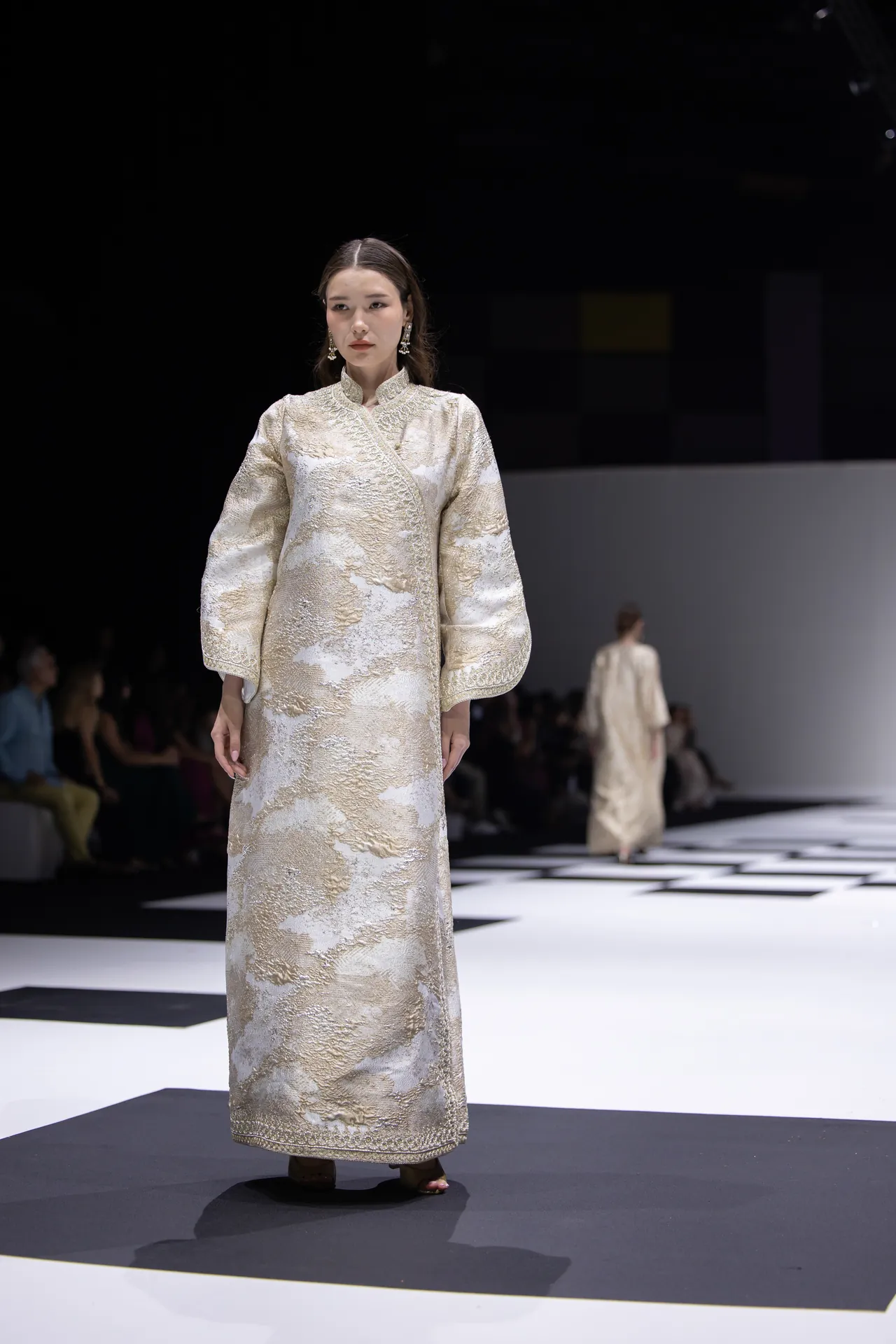 Model walks a runway wearing a long, cream-gold brocade gown with puffed sleeves during a fashion show. The ornate garment features metallic floral patterns and a high collar, with a checkerboard stage beneath her.