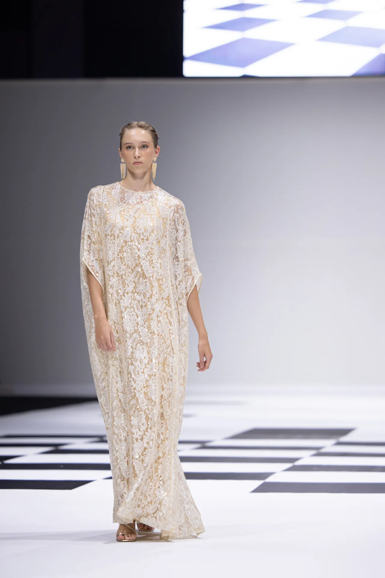 Model walking a fashion runway in a cream lace gown with gold floral embroidery, oversized statement earrings, and low-heeled sandals on a black-and-white checkered catwalk.