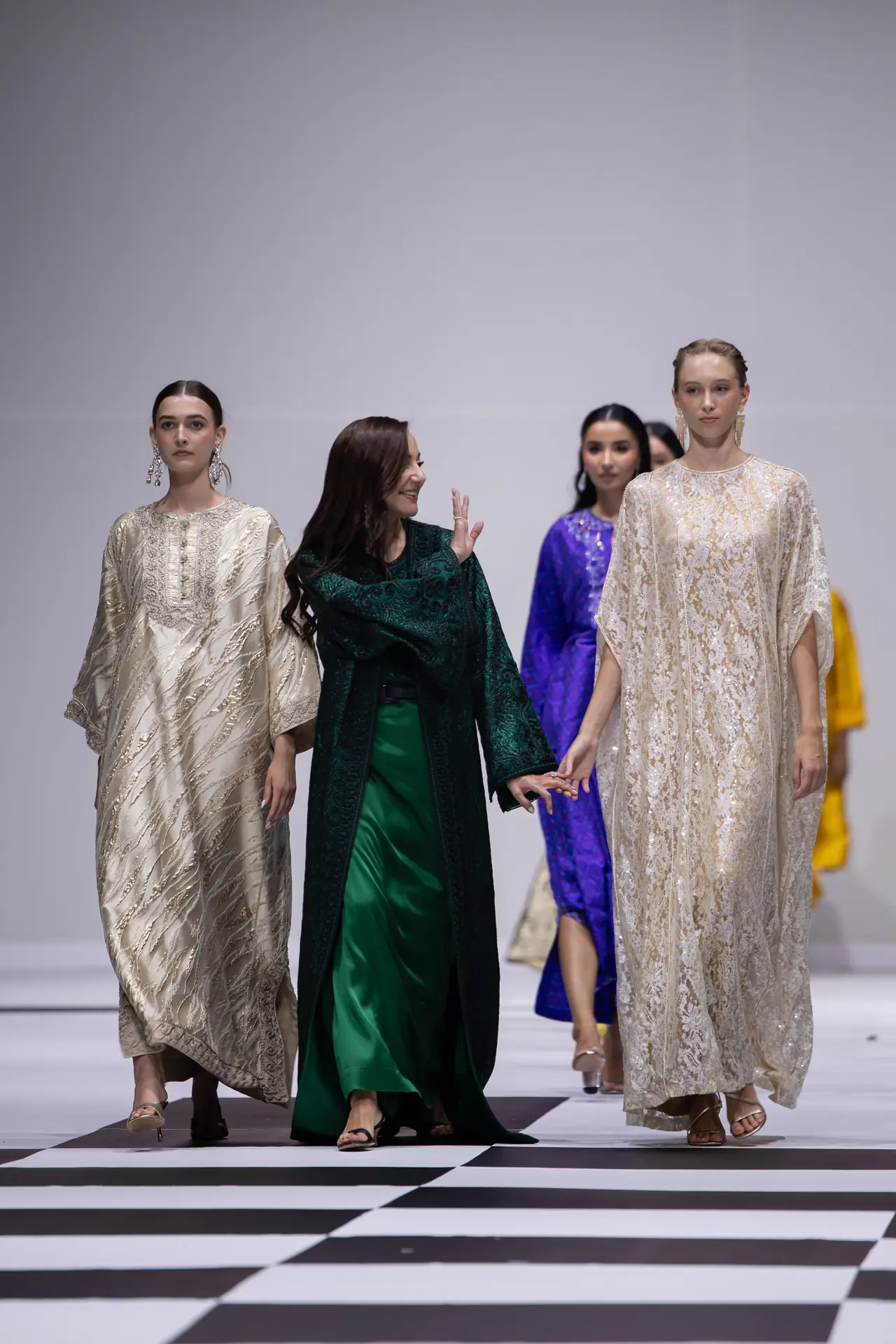 Models walk a fashion show runway in elegant gowns, with a foreground figure in a dark green outfit flanked by two beige lace dresses.