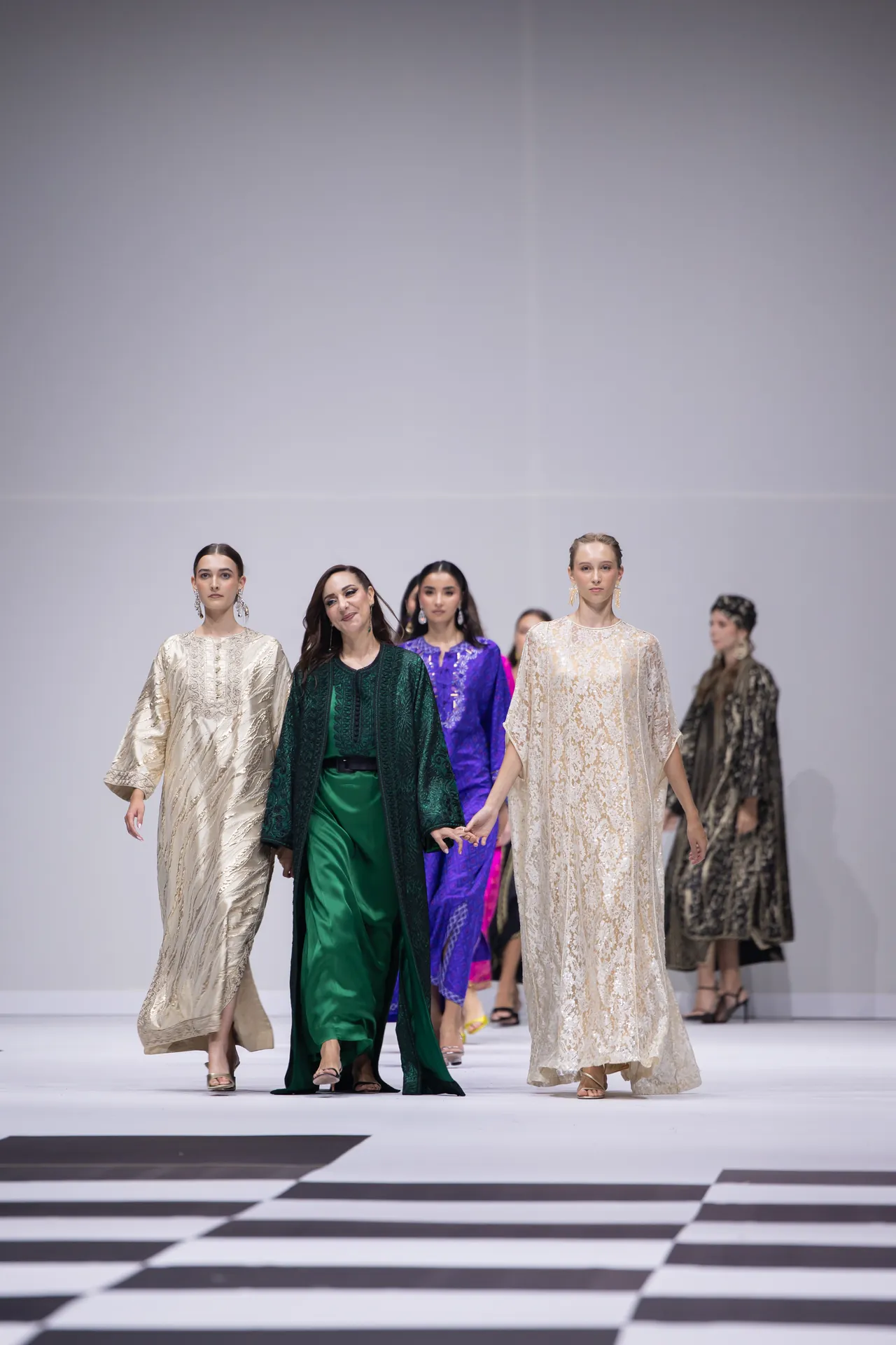 Models walk a fashion runway in flowing, patterned gowns in ivory, green, and purple, with bold striped floor beneath them.