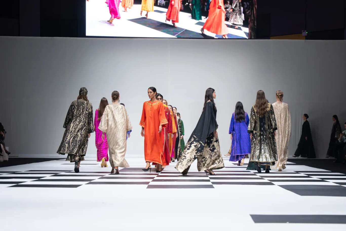 Models in colorful, flowing gowns walk down a black-and-white checkered runway during a fashion show.