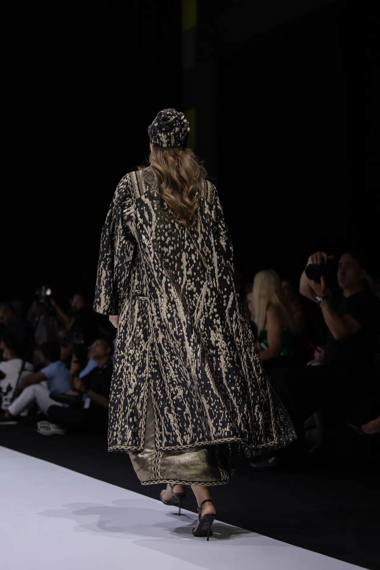 Model seen from behind walking a runway in a black-and-gold embroidered coat and matching hat; audience and photographers in background.
