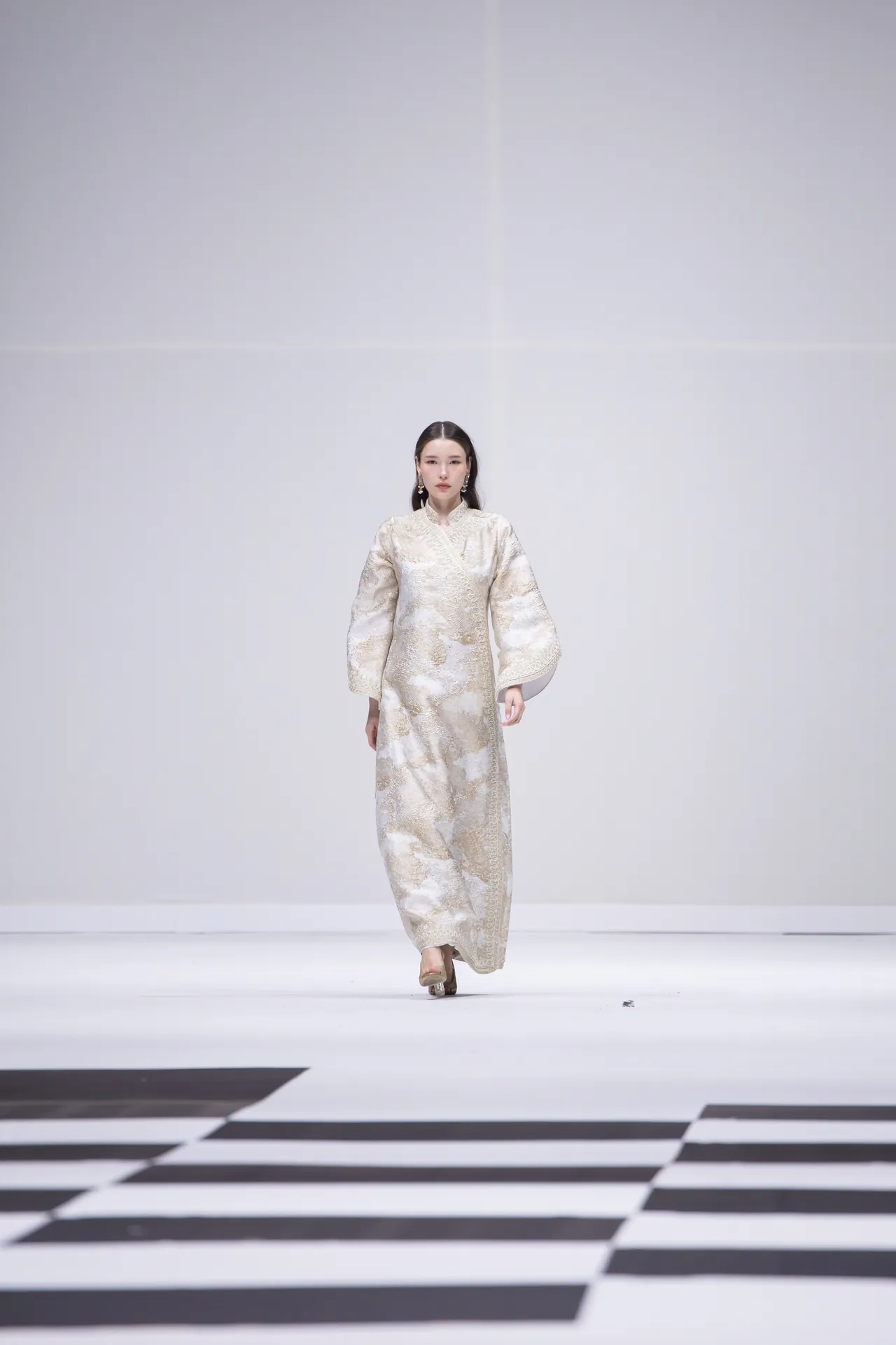 Model walking on a fashion show runway wearing a long cream and gold patterned robe-style dress on a white stage. Padding of black-and-white border visible at the foreground.