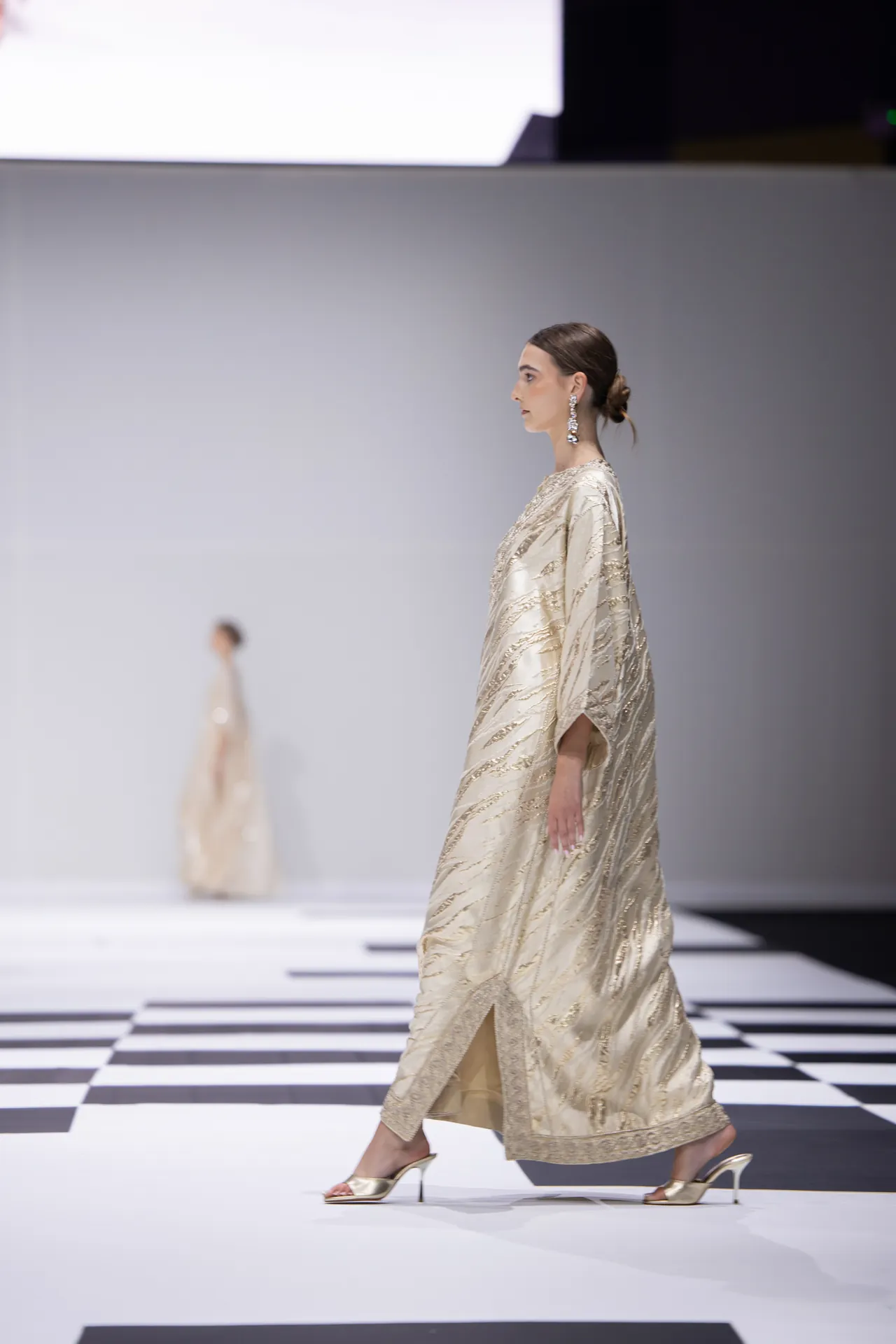 Model walks the runway in a gold-embellished beige gown, side profile, wearing dangling earrings on a black-and-white checkered floor.