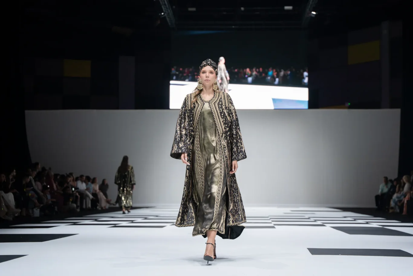 Model walking the runway in a gold-and-black embroidered robe with a matching headscarf; checkerboard catwalk and seated audience in the background.