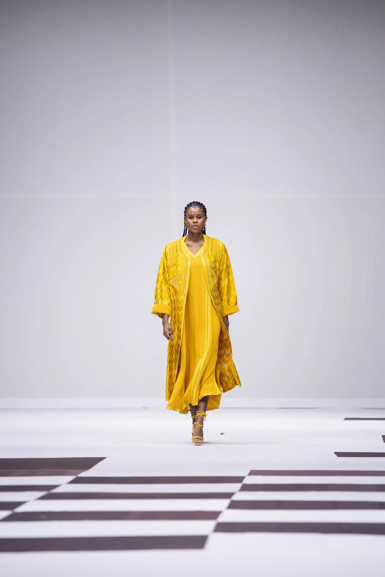 Model walks a fashion runway in a bright yellow, flowing dress with a matching patterned yellow coat; she wears yellow strappy heels, and a plain gray backdrop behind.
