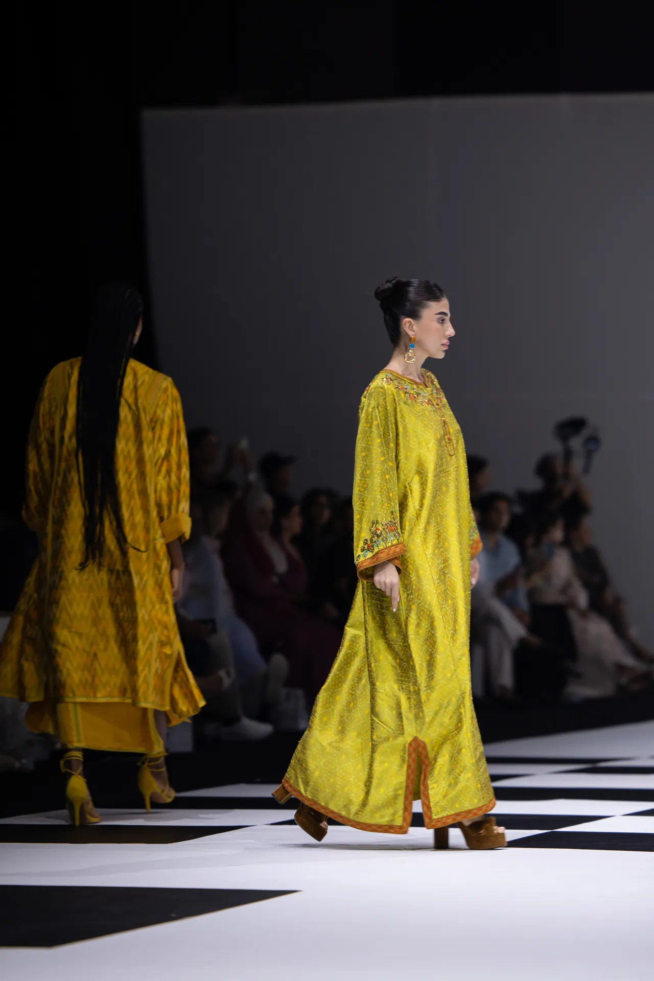Model in a bright yellow, embroidered gown walks the runway, audience in soft focus in the background.