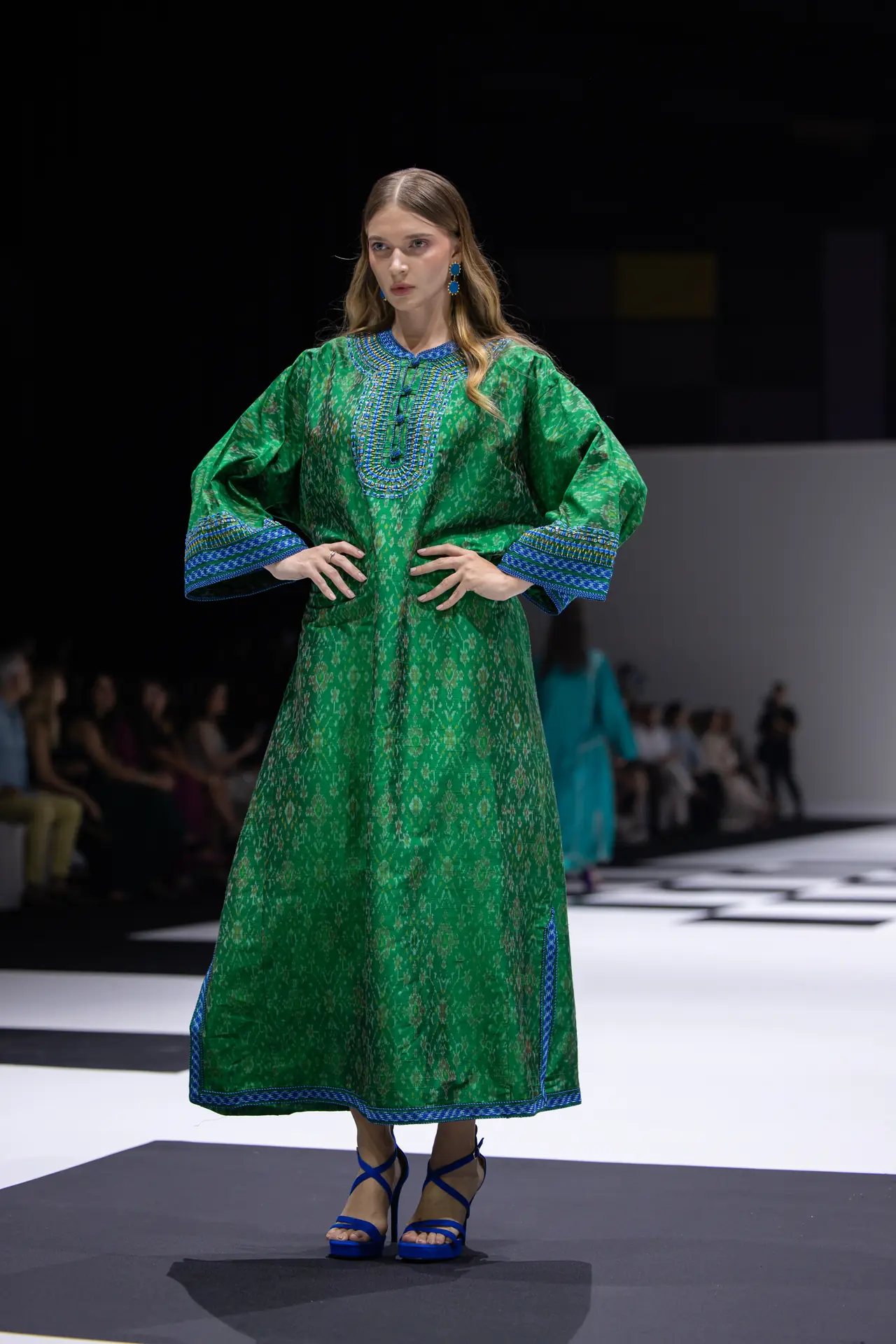 Female model on a runway wearing a green patterned kaftan with blue embroidery and matching blue sandals.