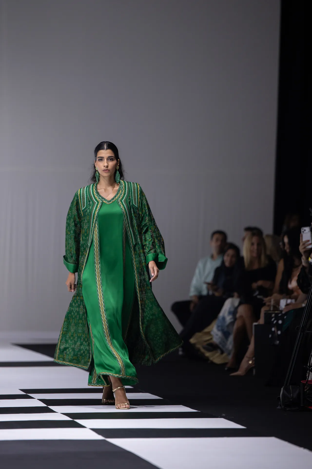 Model walks the runway in a bright green, patterned cape over a green dress; audience seated along the catwalk.