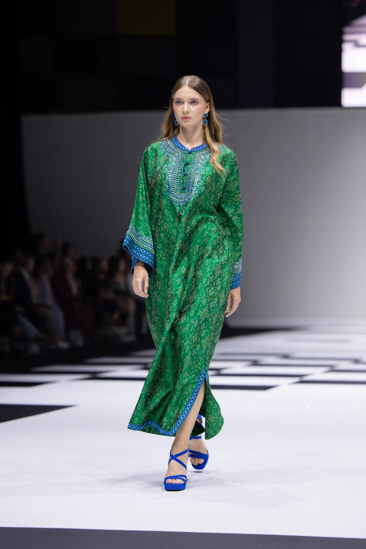 Model on a runway wearing a green printed kaftan with blue embroidery and blue sandals.