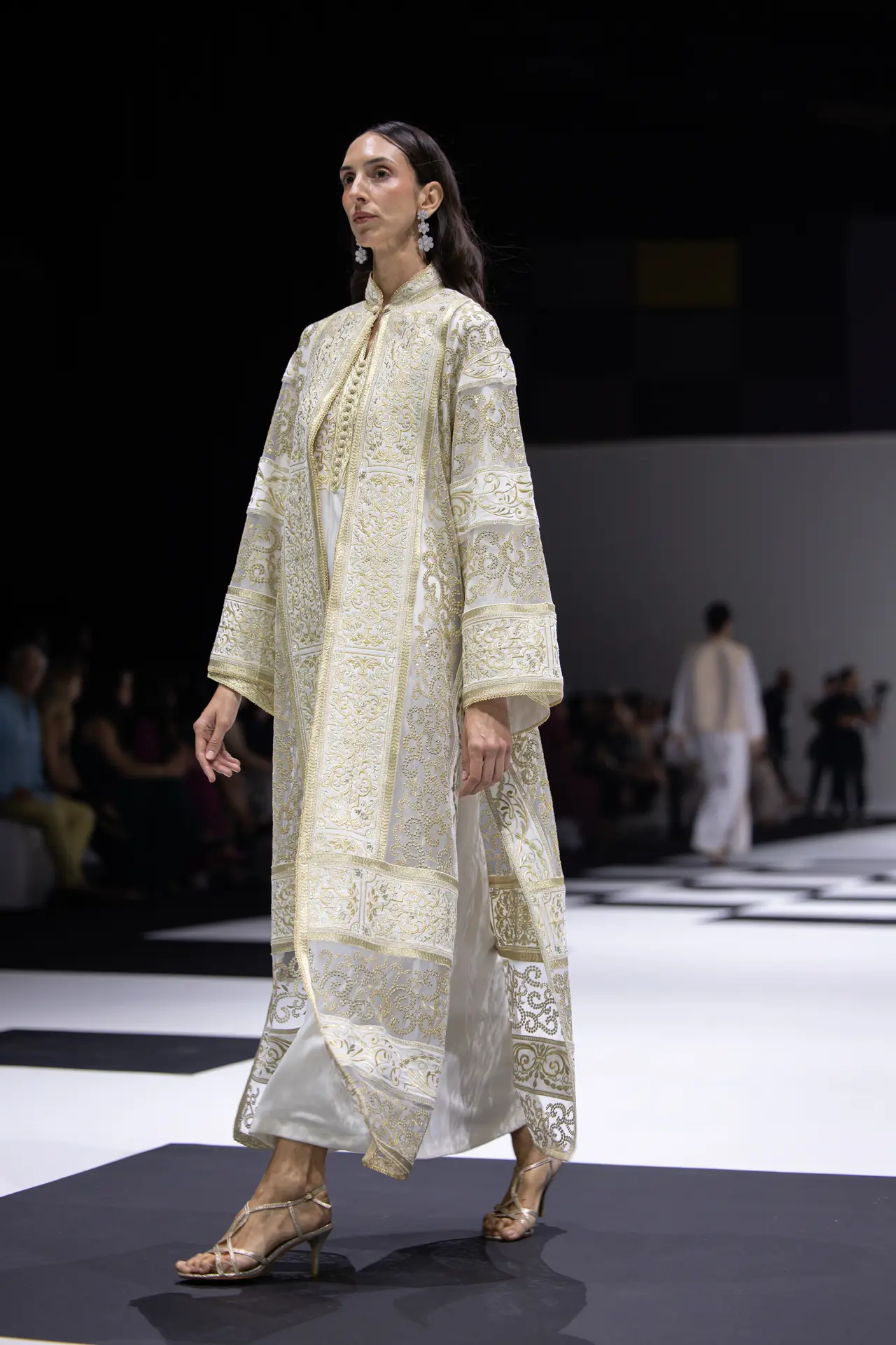 Model walking a fashion runway in an ivory-gold, heavily embroidered long coat with matching white pants, strappy heels.