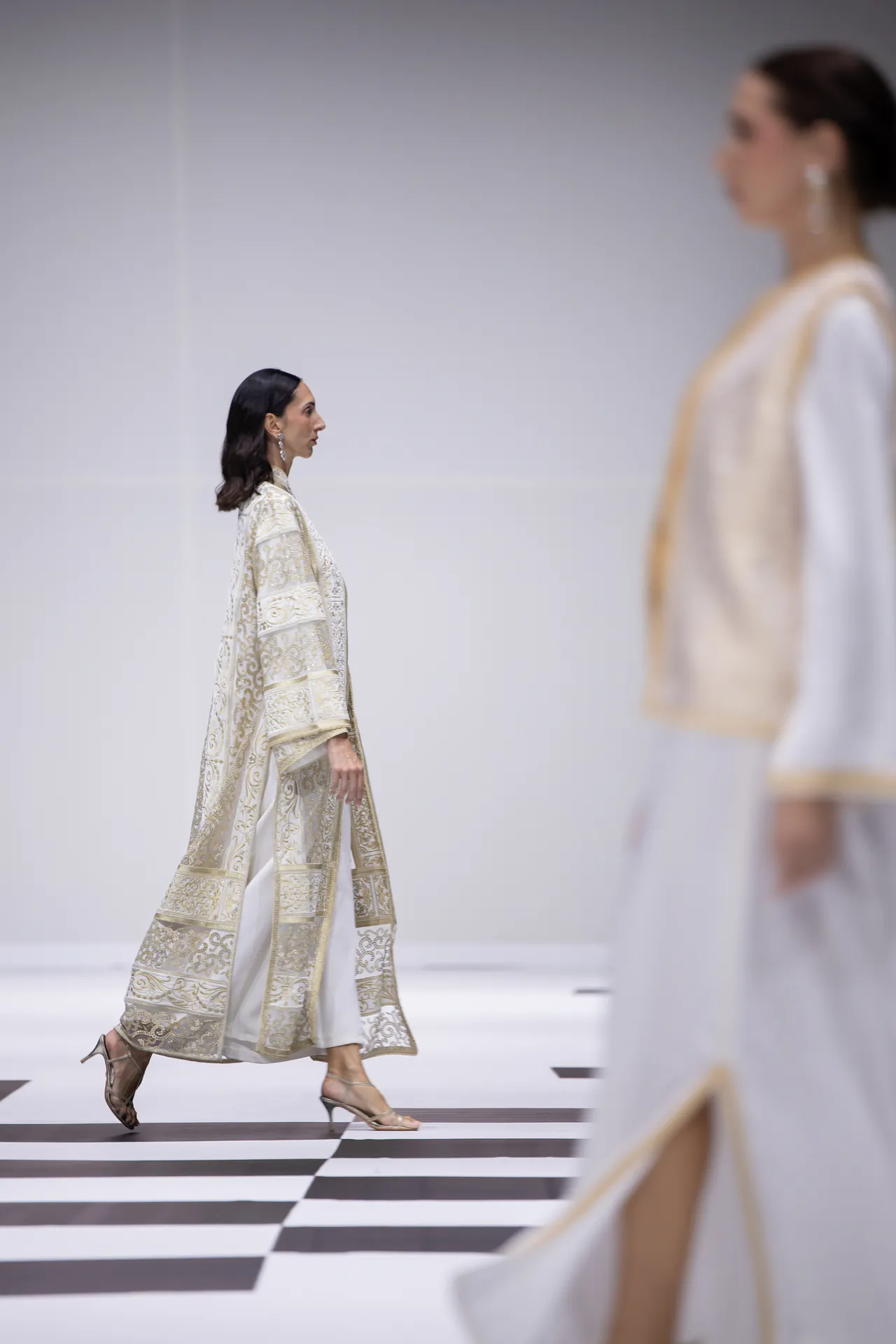 Model walking the runway in a cream and gold embroidered coat with white pants; foreground model blurred in the foreground.