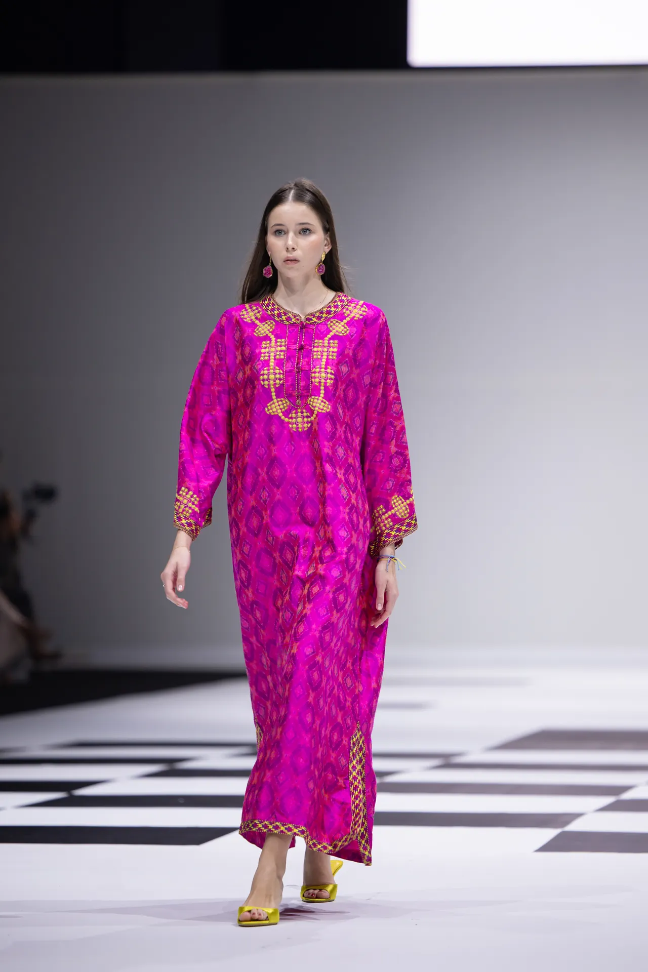 Model walks the runway in a bright magenta traditional dress with gold embroidery and geometric patterns, complemented by pink earrings and yellow sandals.