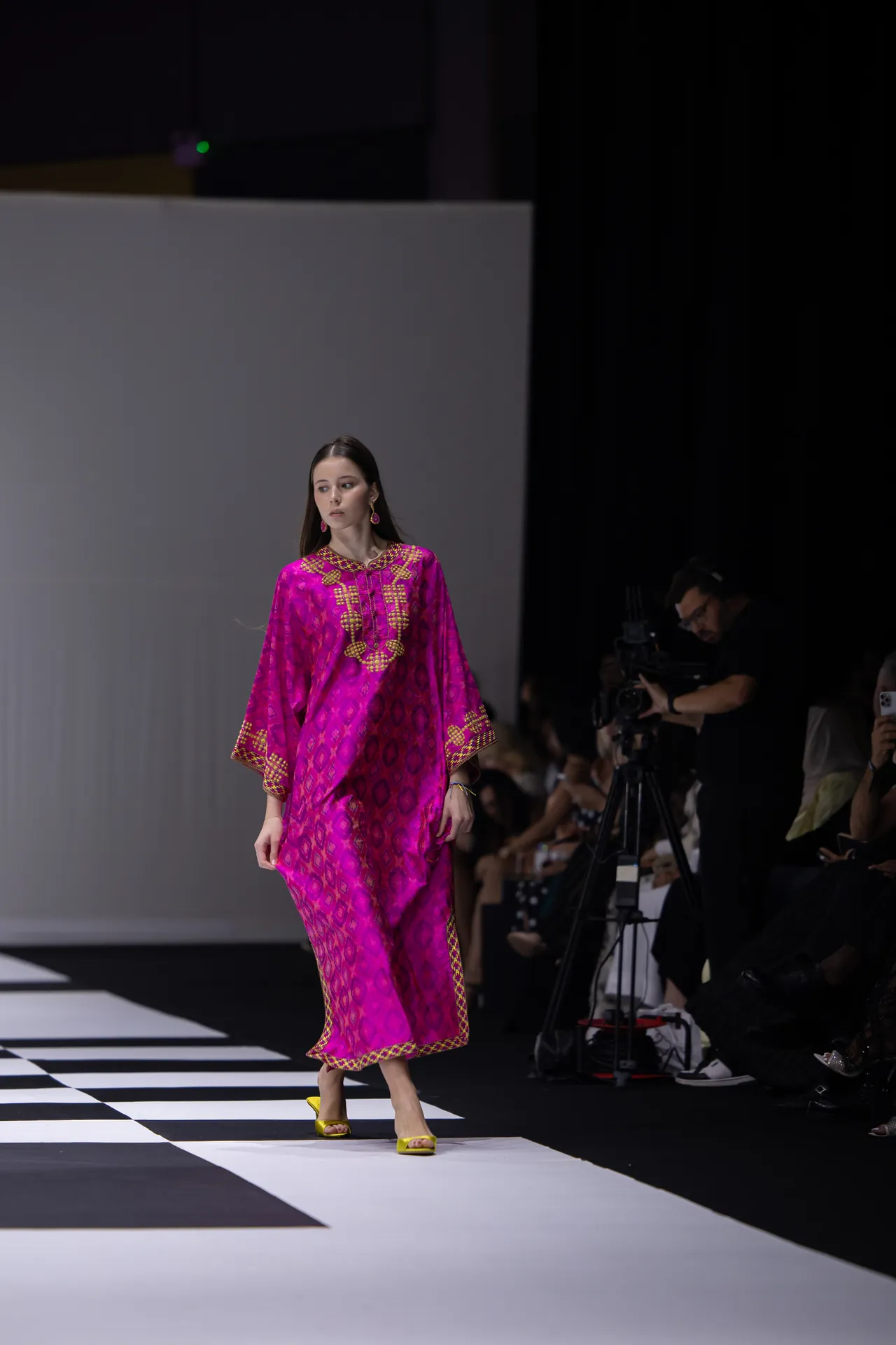 Model walks the runway in a bright pink traditional outfit with gold embroidery and yellow heels, photographers in the background.