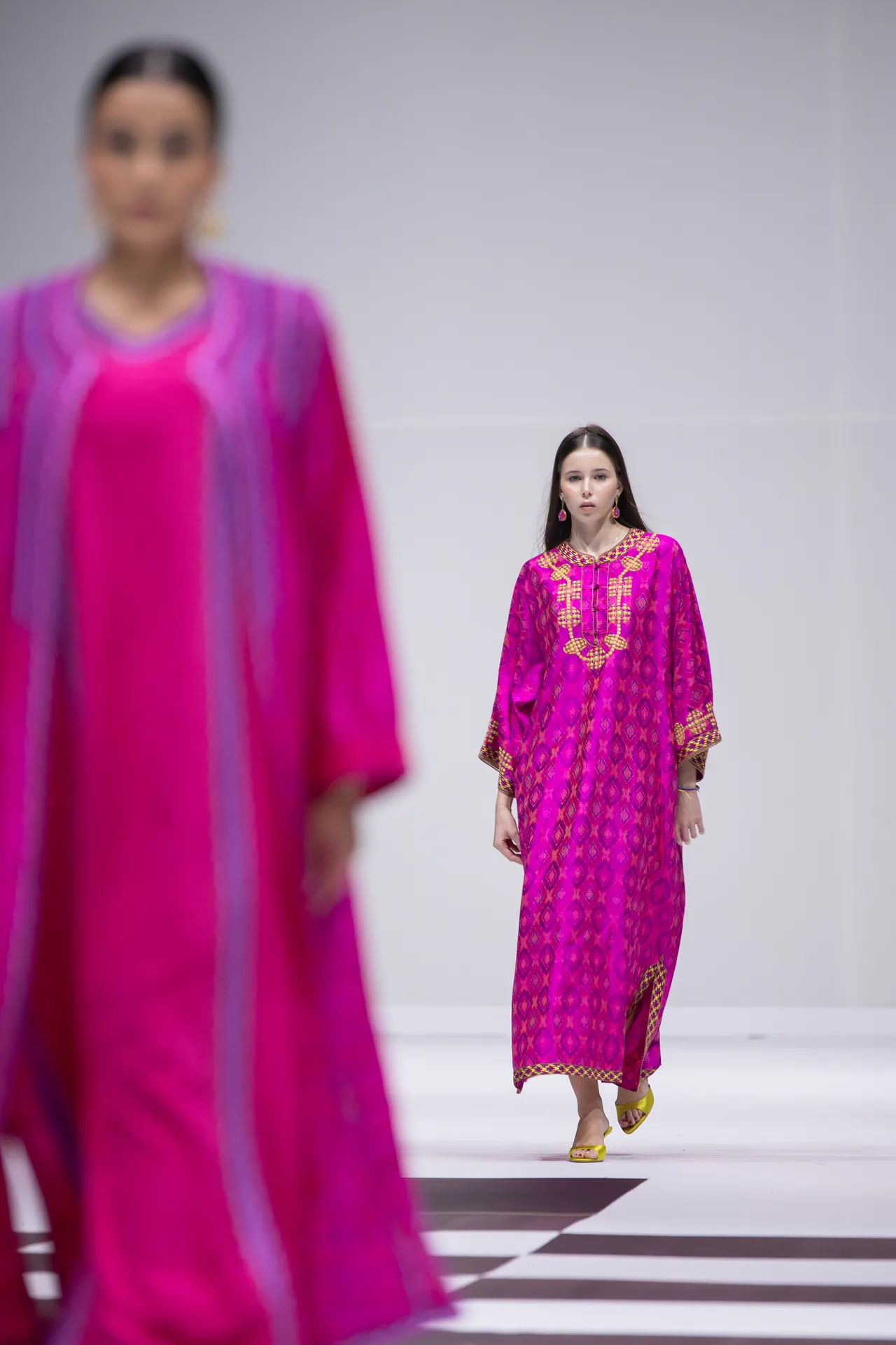 Model walking a runway in a bright magenta printed kaftan with gold embroidery; foreground blur of another pink garment/frame foreground, yellow sandals visible.