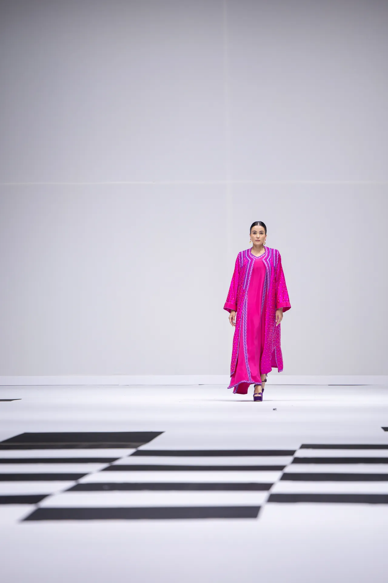 Model wearing a bright pink traditional outfit walks down a minimalist white runway with a black-and-white pattern on the floor.
