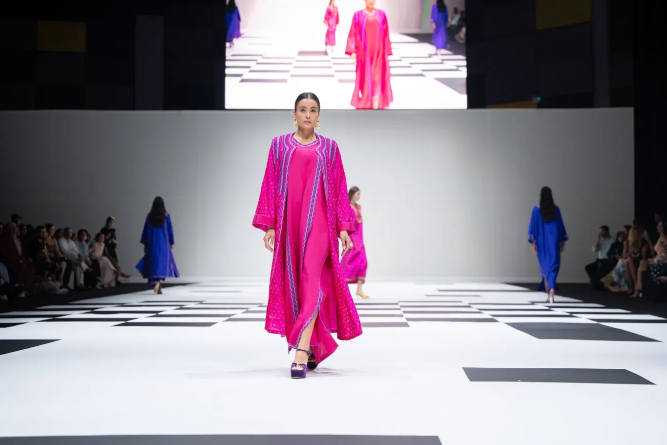 Model walks the runway in a bright pink dress with a patterned cape as spectators watch from the sides.