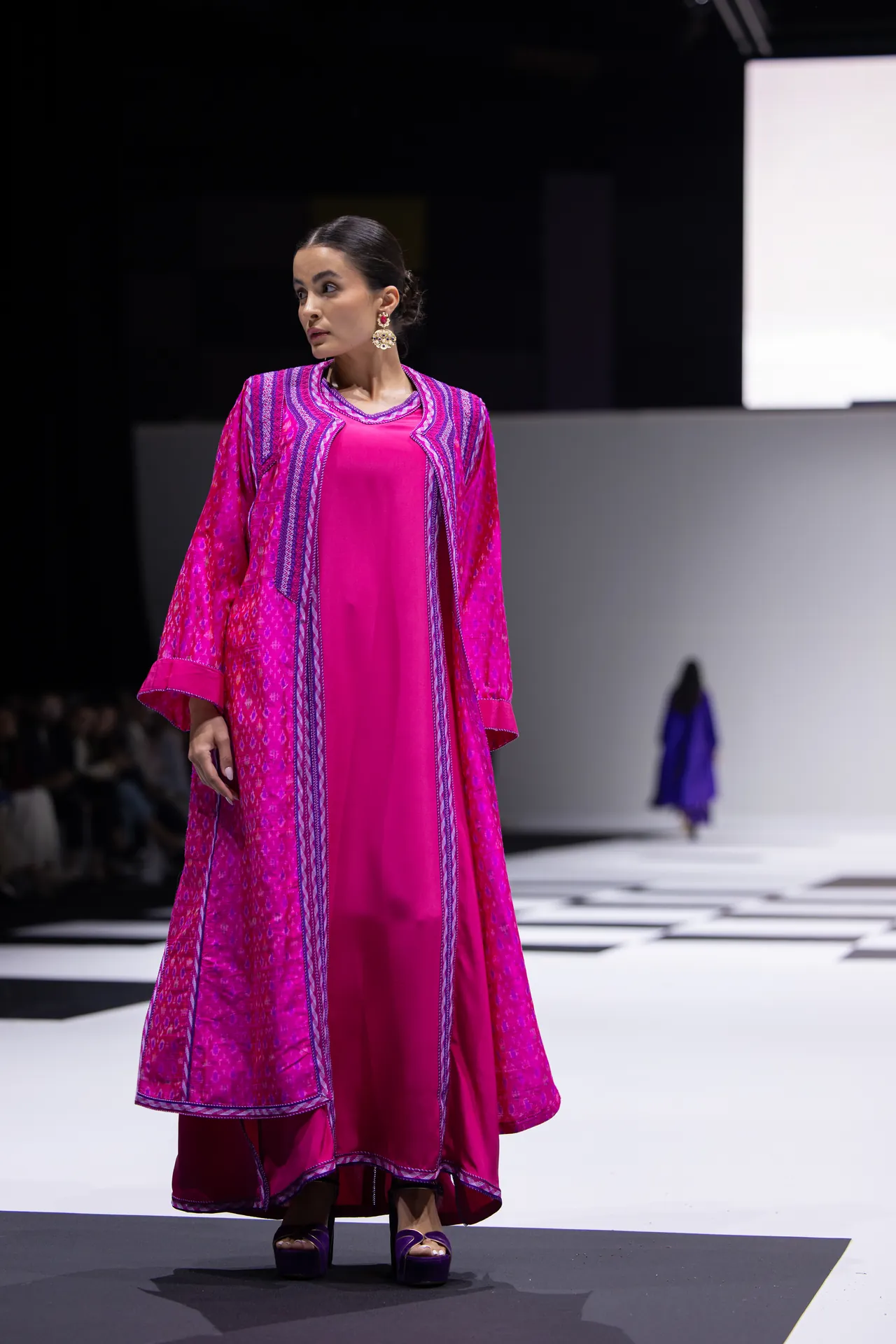 Model walking a fashion runway in a bright pink traditional dress with purple-embroidered cape and statement earrings on a checkered catwalk.