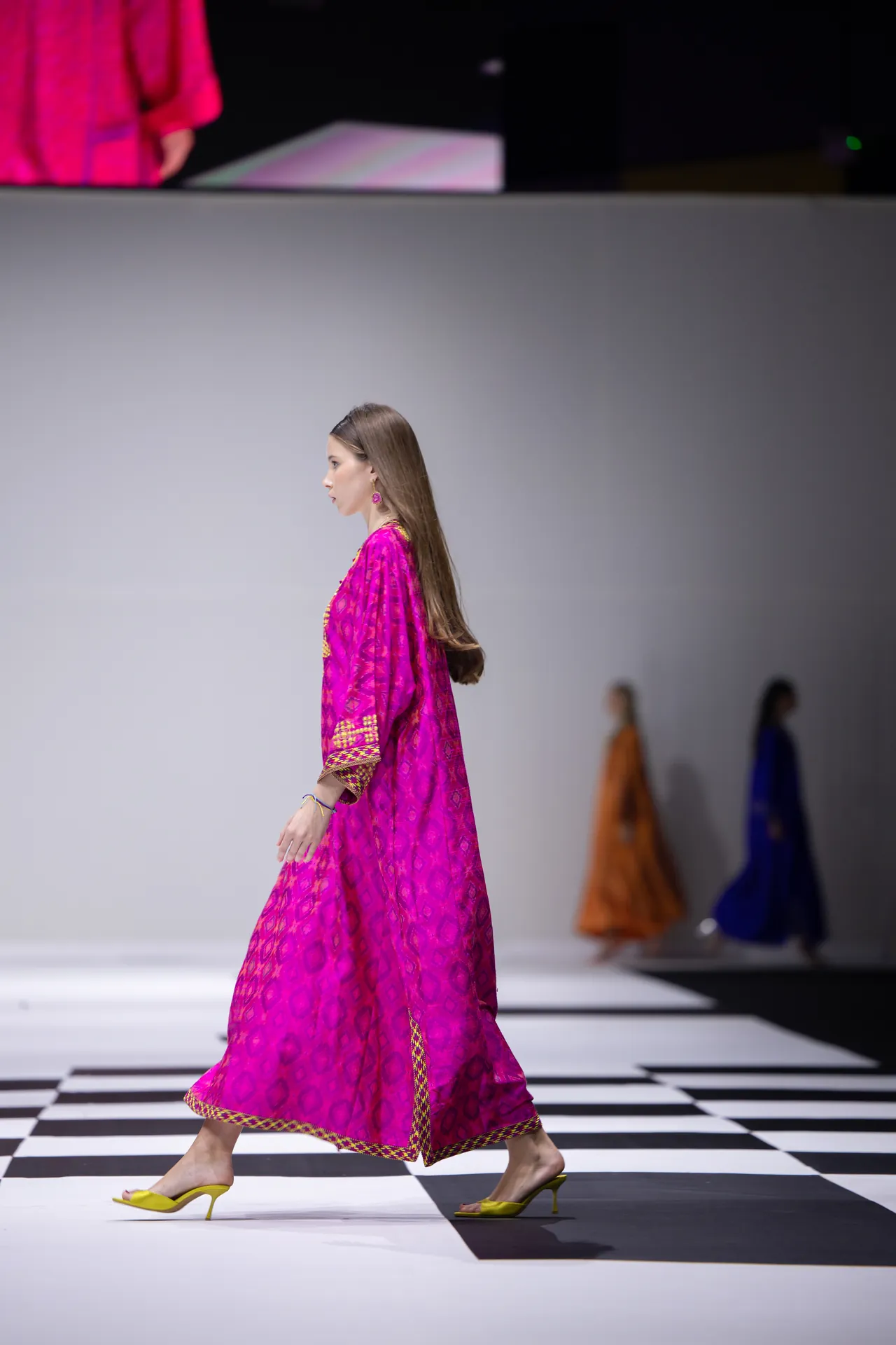 Model walking a runway in a bright pink patterned dress with yellow heels on a black-and-white checkered catwalk.