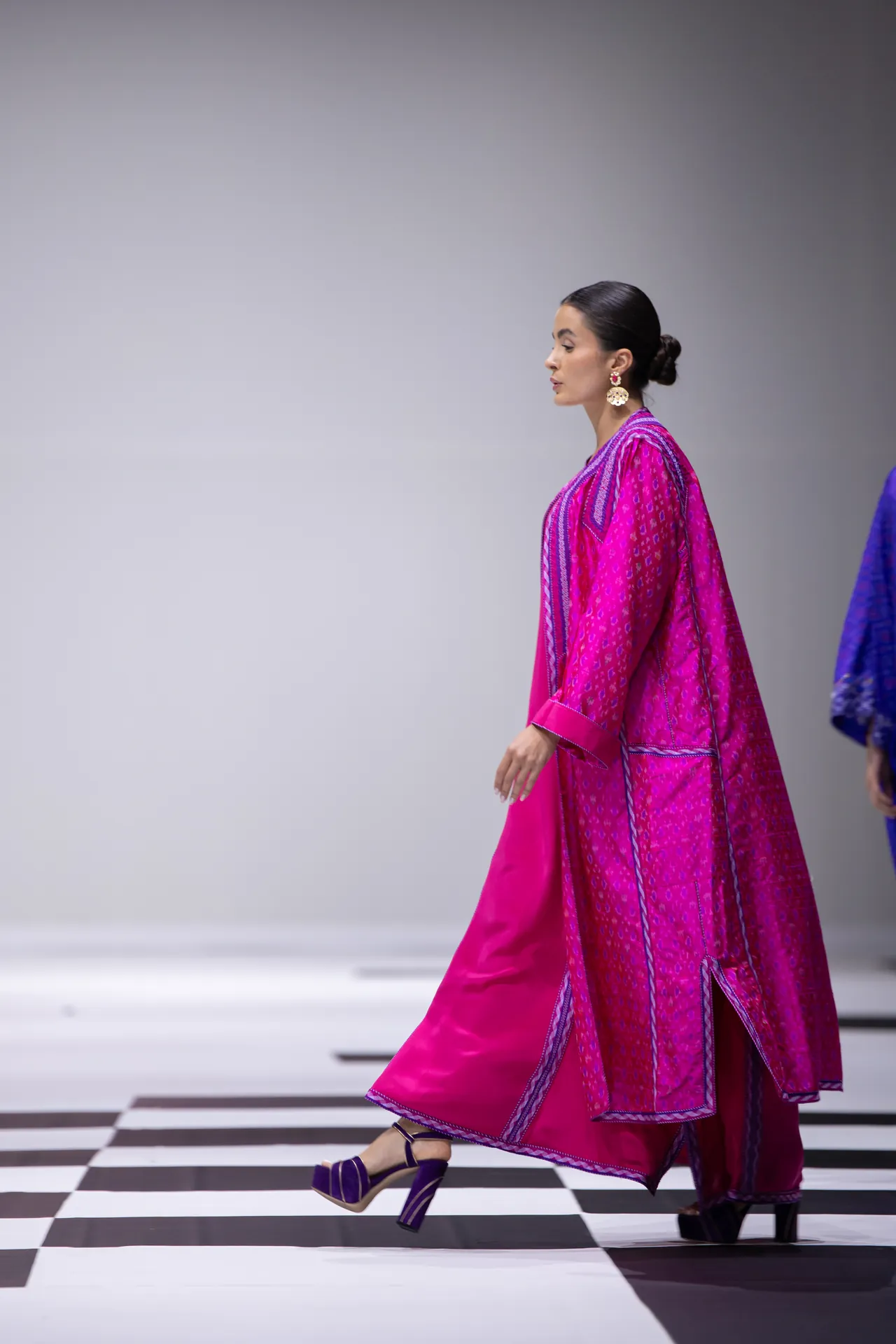 Model in a vivid pink traditional outfit walking the runway, with a checkered floor beneath her.