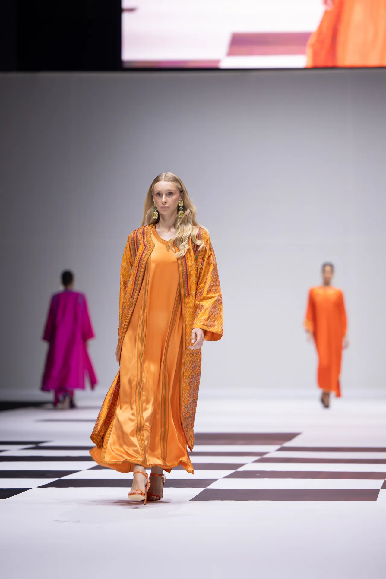 Model walks the runway in an orange satin dress with a patterned cape and bold earrings on a black-and-white checkered floor runway.