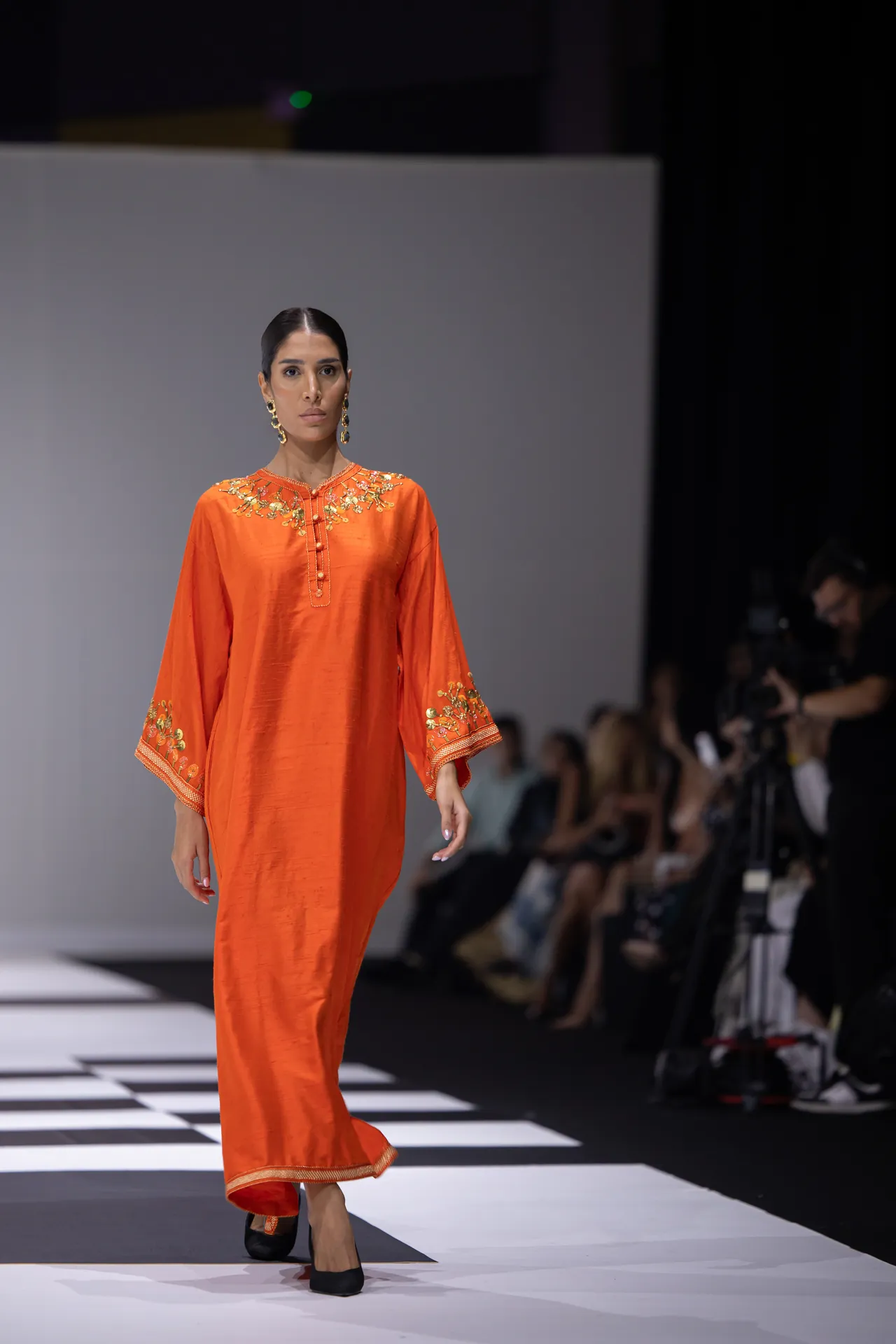Model walking a runway in an orange dress with gold embroidery at the neckline and cuffs.