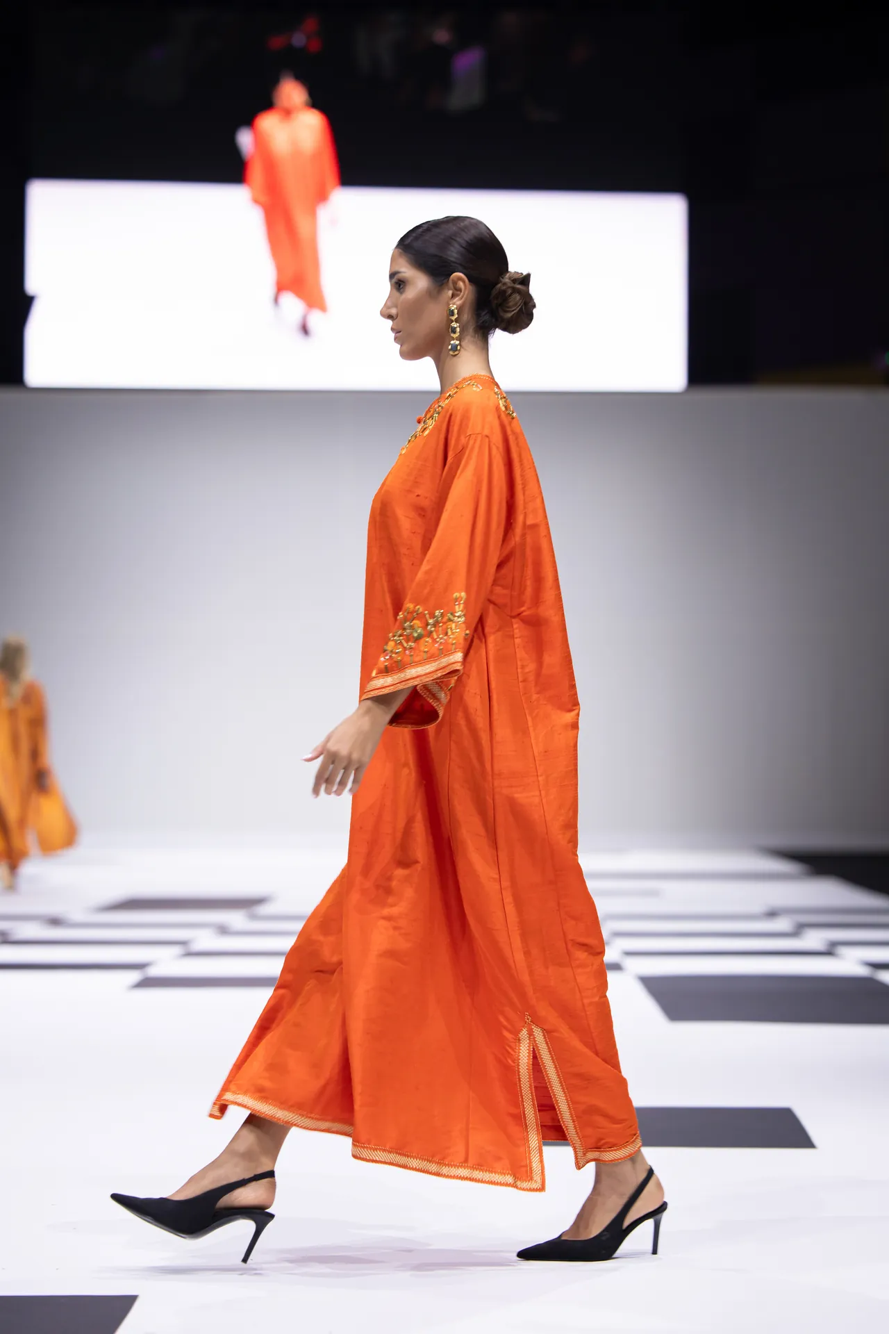 Model walking sideways on a fashion runway in an orange embroidered dress, black heels, hair in a bun, white runway with black checkered floor pattern in the background.