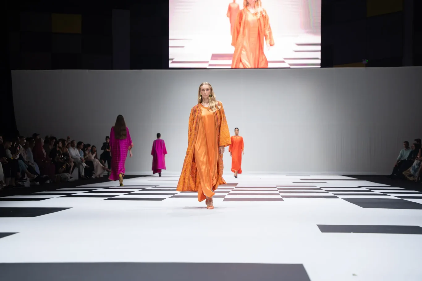 Model walking the runway in an orange flowing outfit with a matching cape; checkerboard runway, audience on both sides, large screen backdrop.