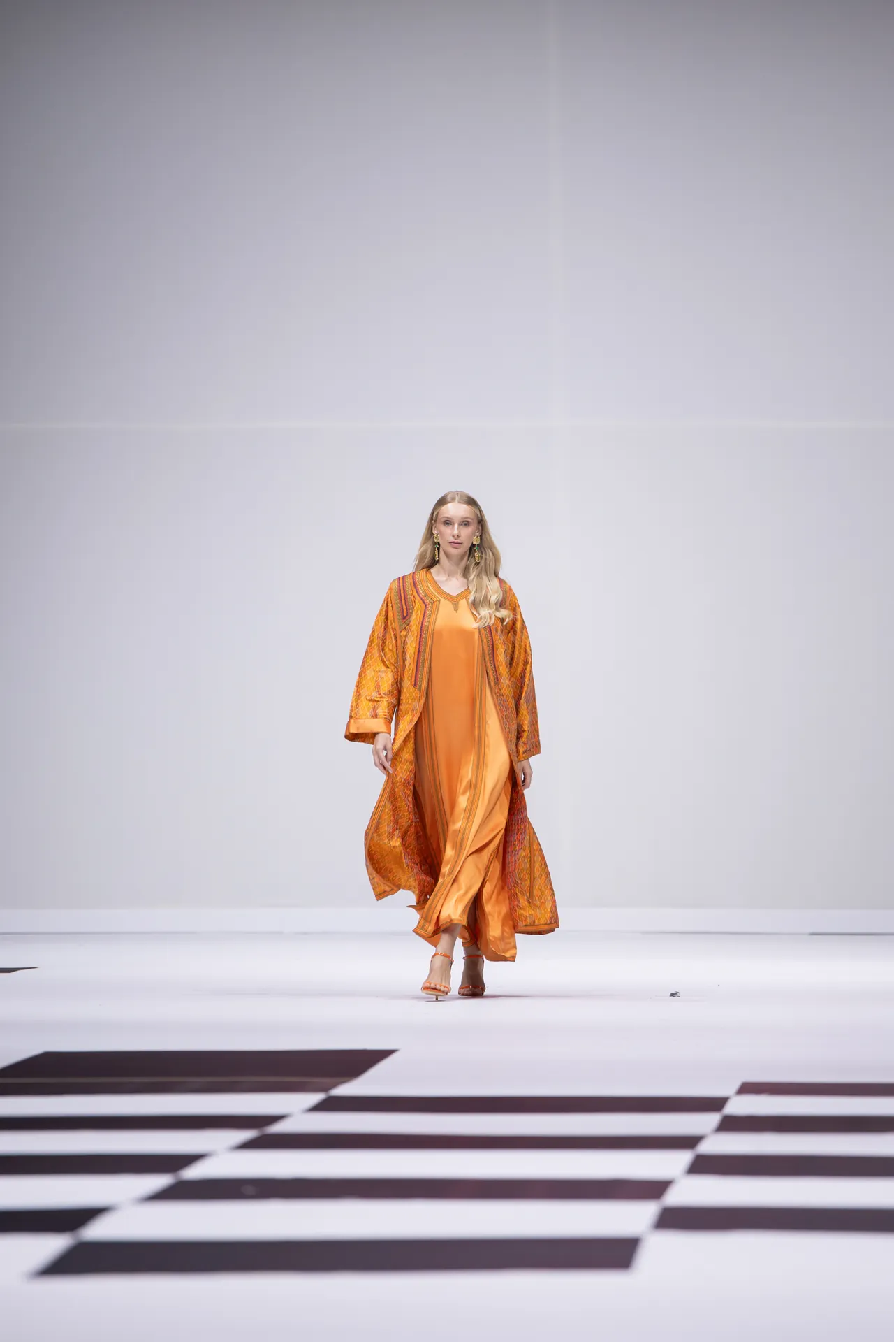 Model walking the runway in a flowing orange dress and patterned shawl, against a minimalist backdrop, orange heels visible