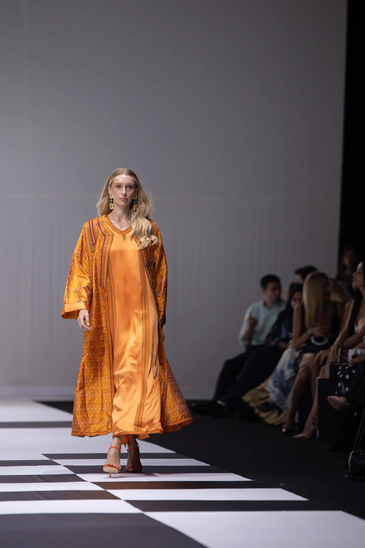 Model walks the runway in an orange satin dress with a patterned orange cape, while spectators watch from the sides.