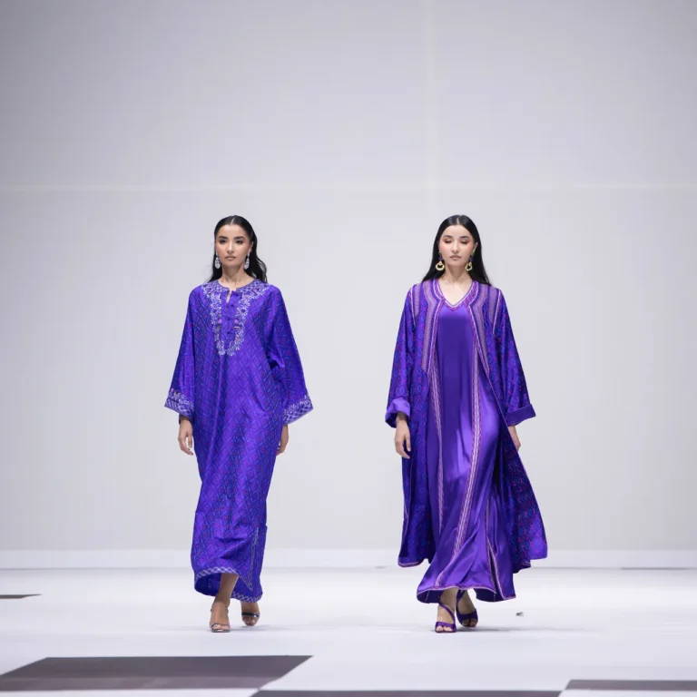 Two models in purple outfits walk the runway; left in a patterned purple kaftan, right in a silky purple gown with a cape-like outer layer.