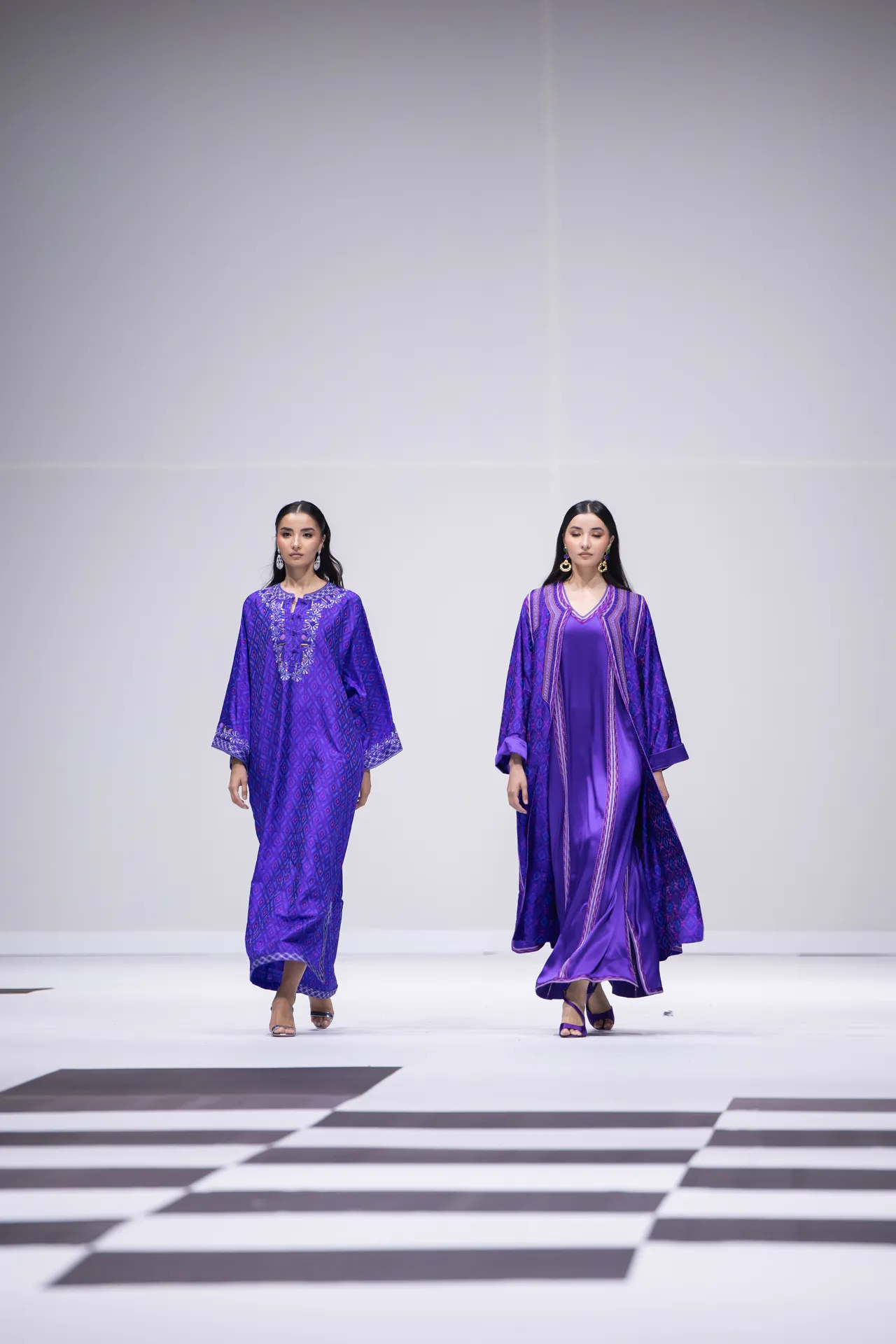 Two models in purple outfits walk the runway; left in a patterned purple kaftan, right in a silky purple gown with a cape-like outer layer.