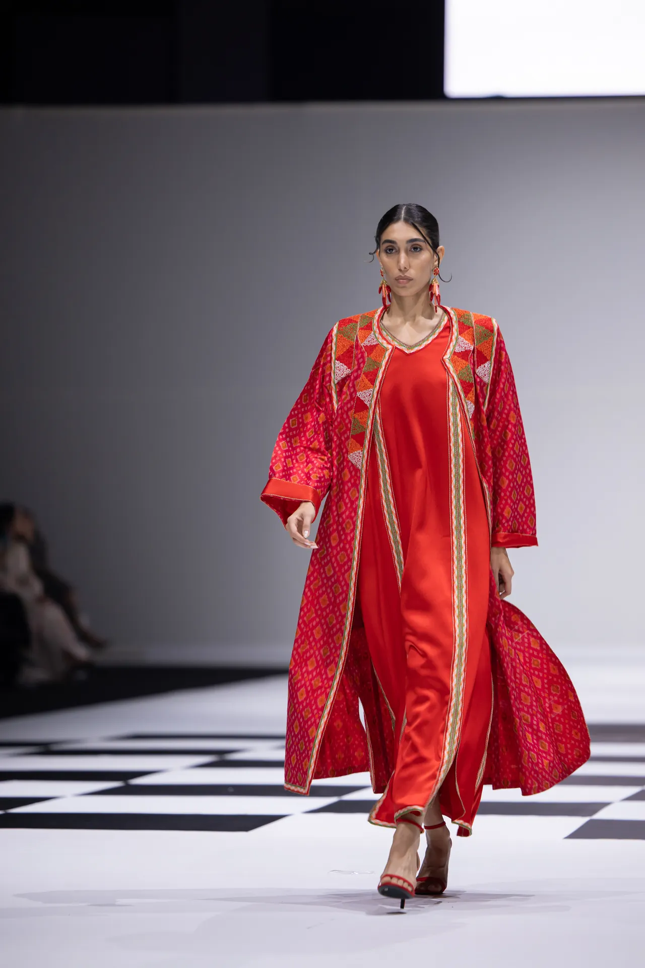 Model walking a fashion runway in a bright red, patterned outer coat over a red satin dress with gold trim; black-and-white checkered floor.