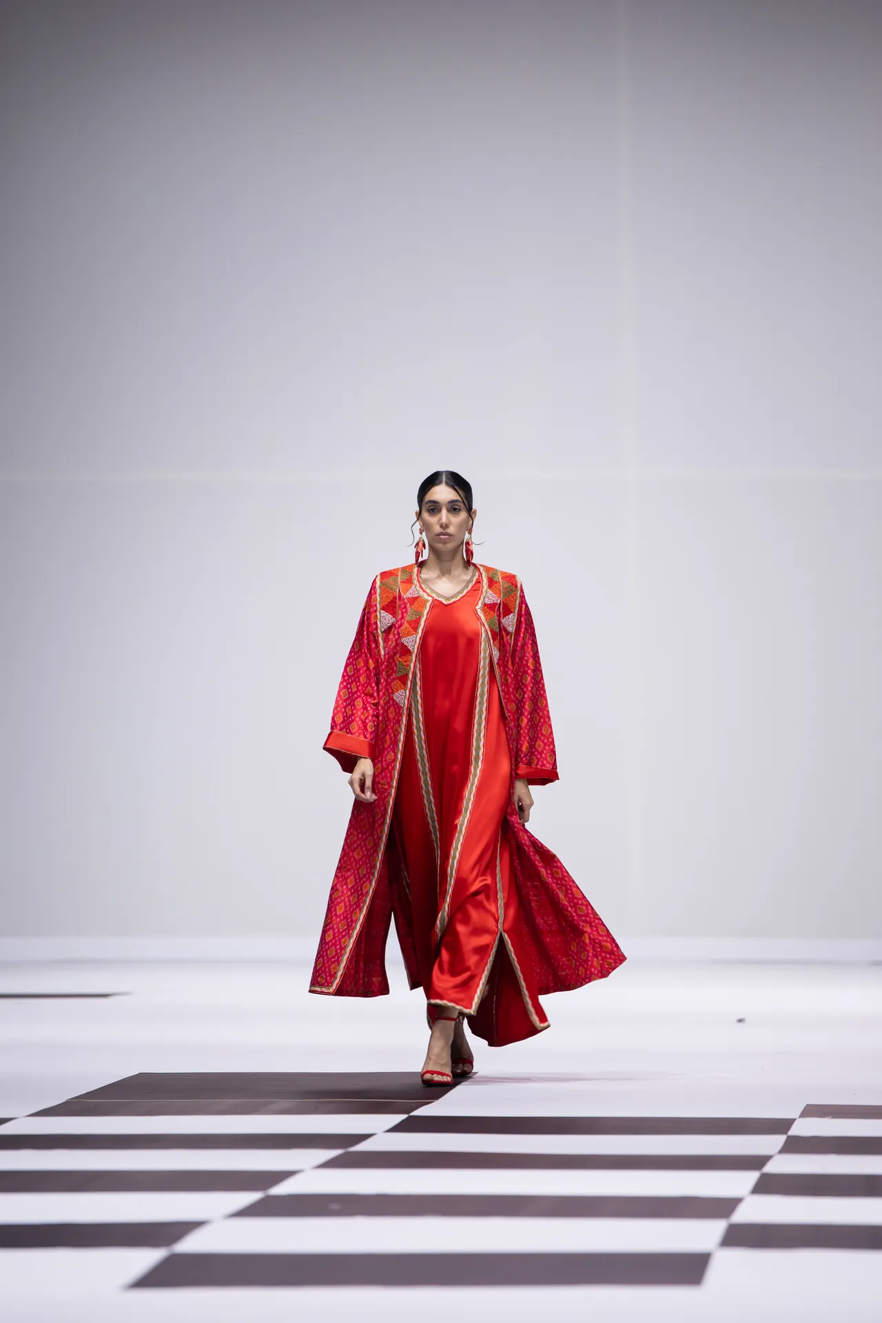 Female model walking runway in a red satin dress with a patterned red cape featuring gold trim on a geometric floor display of a fashion show.