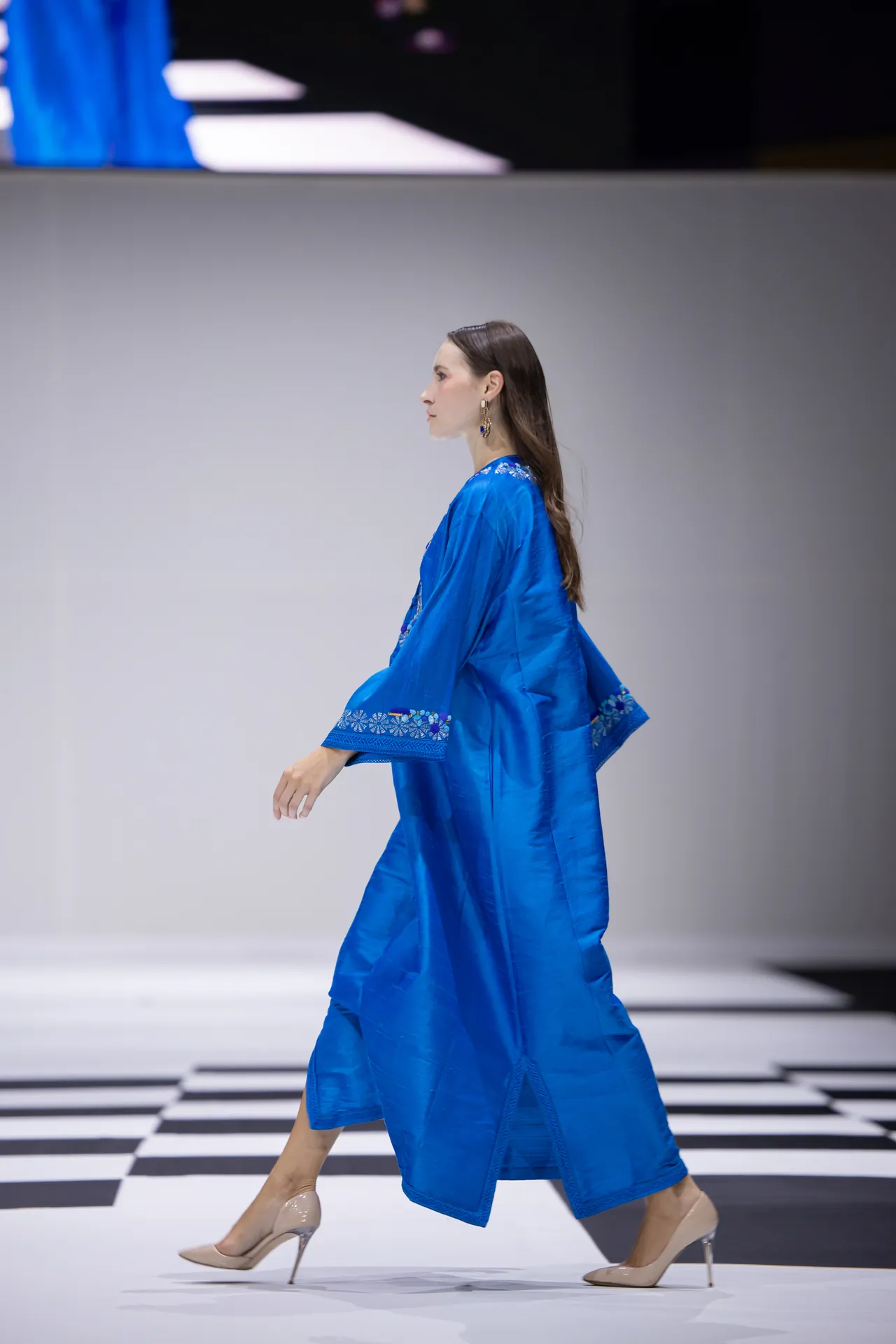 Side profile of a female model walking the runway in a royal blue embroidered robe with beige heels.