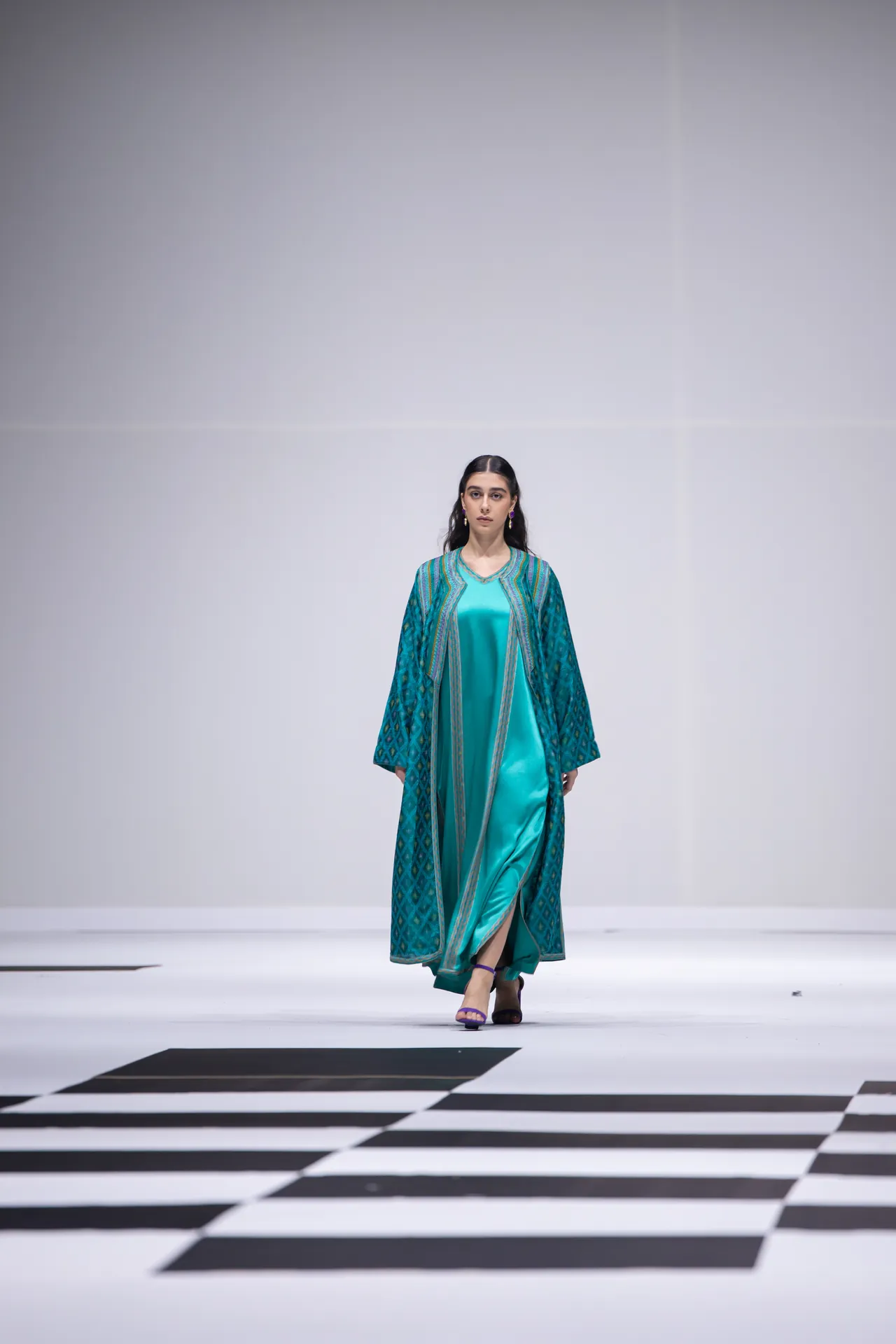 Model walking a runway in a turquoise satin dress with a patterned teal coat, on a black-and-white checkered runway floor.