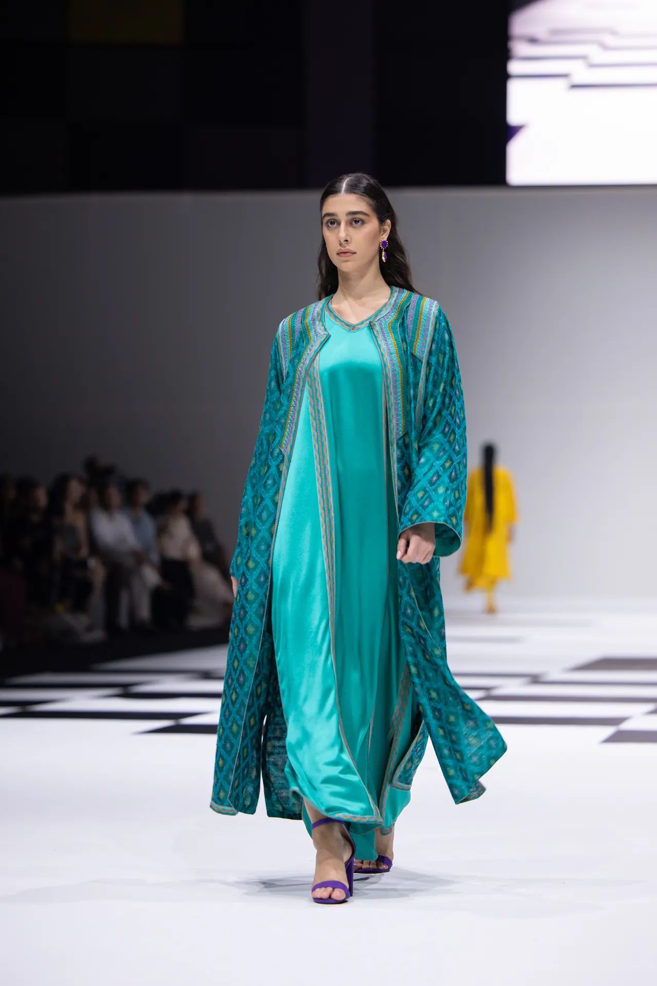 Model walking a runway in a teal satin dress with a blue-green patterned outer coat and purple sandals; audience blurred in the background.