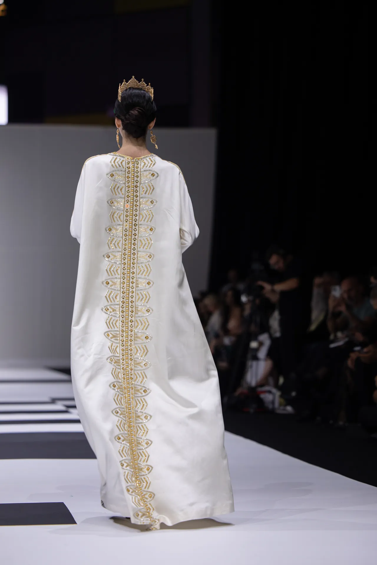 Back view of a model wearing a white gown with gold geometric embroidery and a gold crown on a fashion show runway, audience in the background.