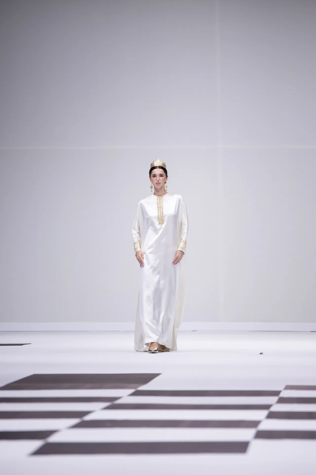 Female model on a runway wearing a long white gown with gold embroidered accents and a gold crown, walking confidenceously.
