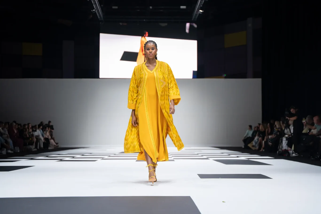 Model walks the runway in a bright yellow geometric-patterned coat over a yellow dress on a black-and-white checkered catwalk.