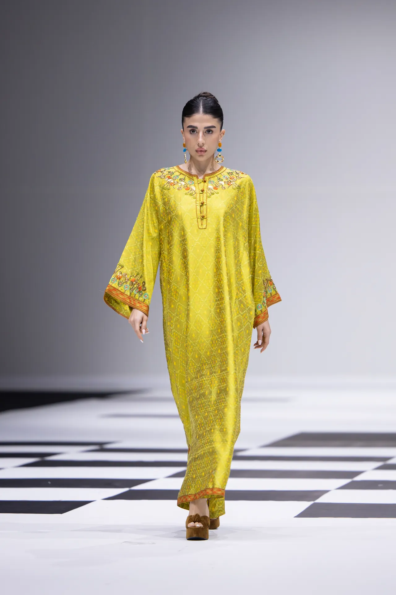 Model walking a runway in a bright yellow embroidered kaftan with orange trim and turquoise earrings, on a black-and-white checkerboard catwalk.