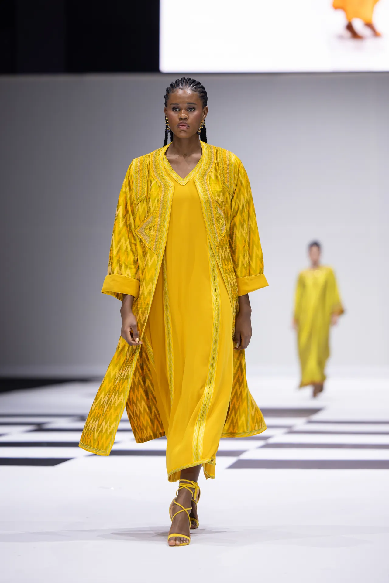 Model walking a runway in a bright yellow embroidered coat over a matching yellow dress, with matching strappy sandals.