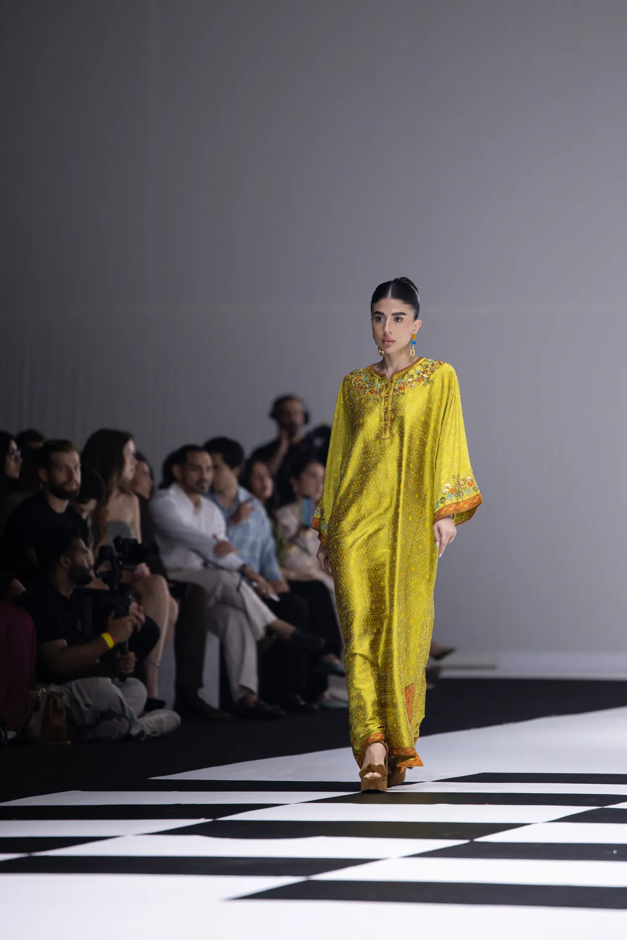 Model in a bright yellow embroidered dress walks the runway as seated audience watches from the sides.