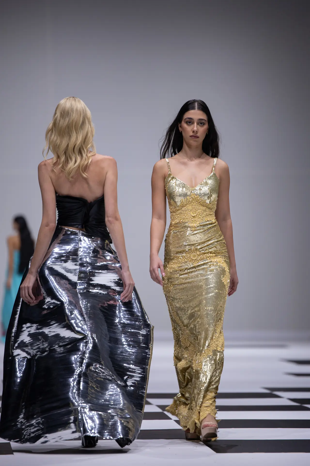 Two models on a fashion runway; one in a gold sequin gown walks toward the camera, the other in a silver metallic skirt is seen from the back.