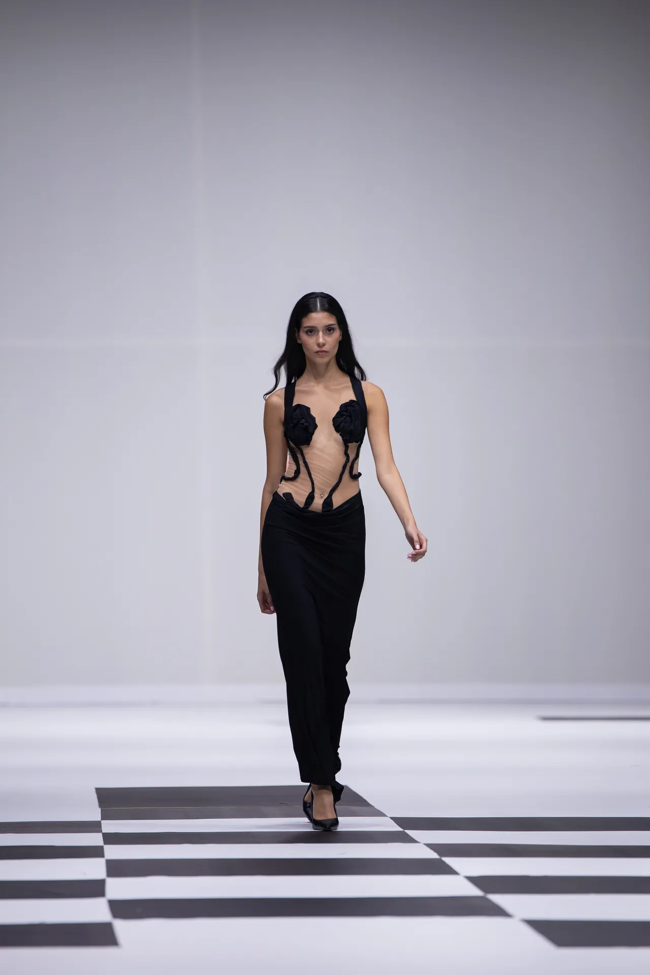 Model walking a fashion show on a white runway, wearing a sheer black cutout top and black pants with a checkerboard floor beneath.