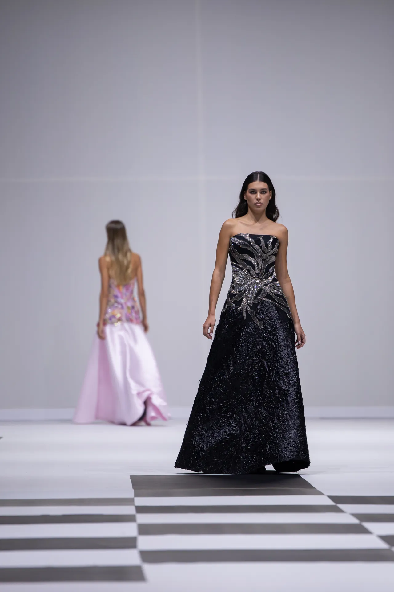 Model on a runway wearing a strapless black gown with silver embellishments; checkered floor and a second model in a pink dress in the background.
