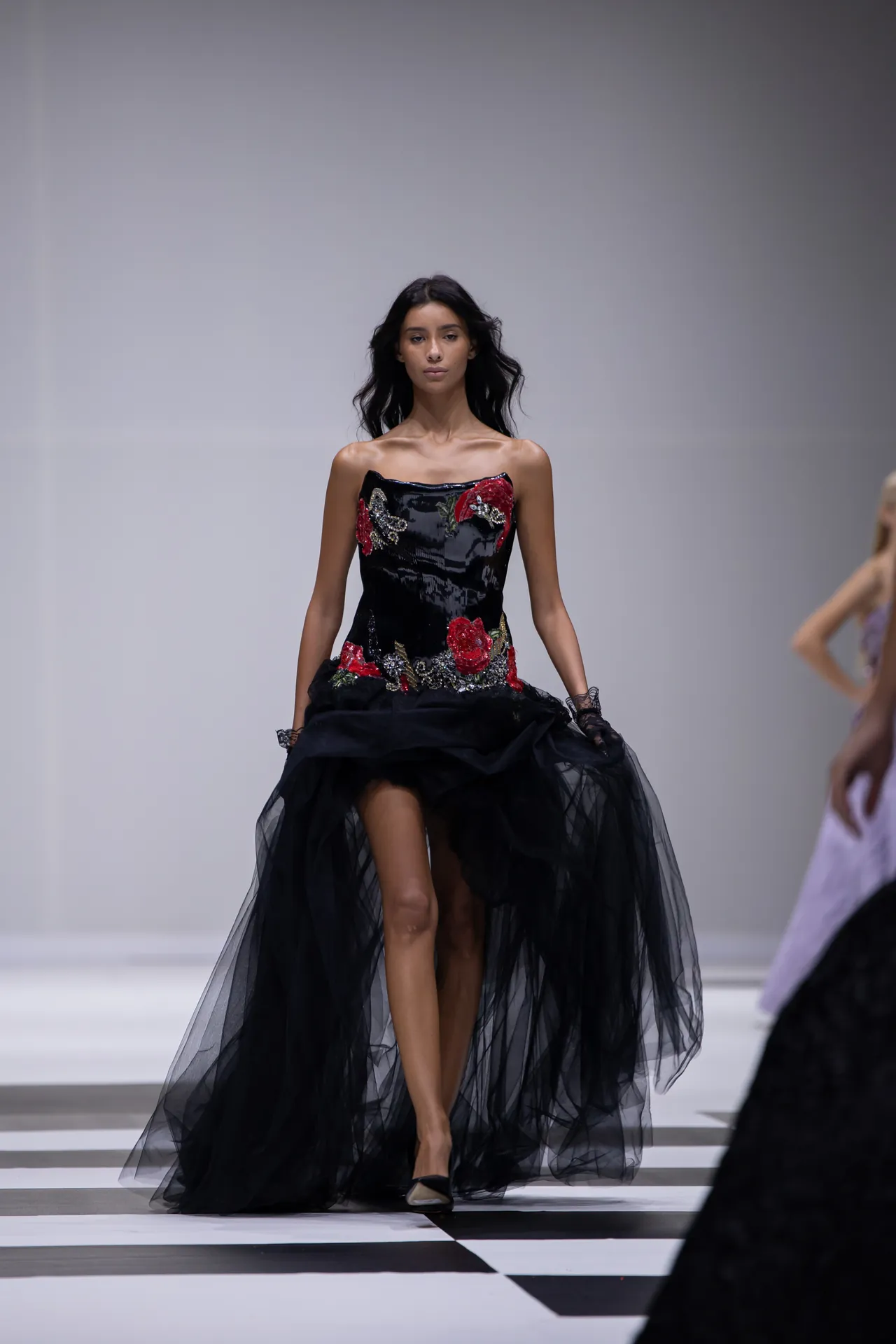 Model walking a fashion runway in a strapless black gown with red floral embroidery and a sheer layered tulle skirt.