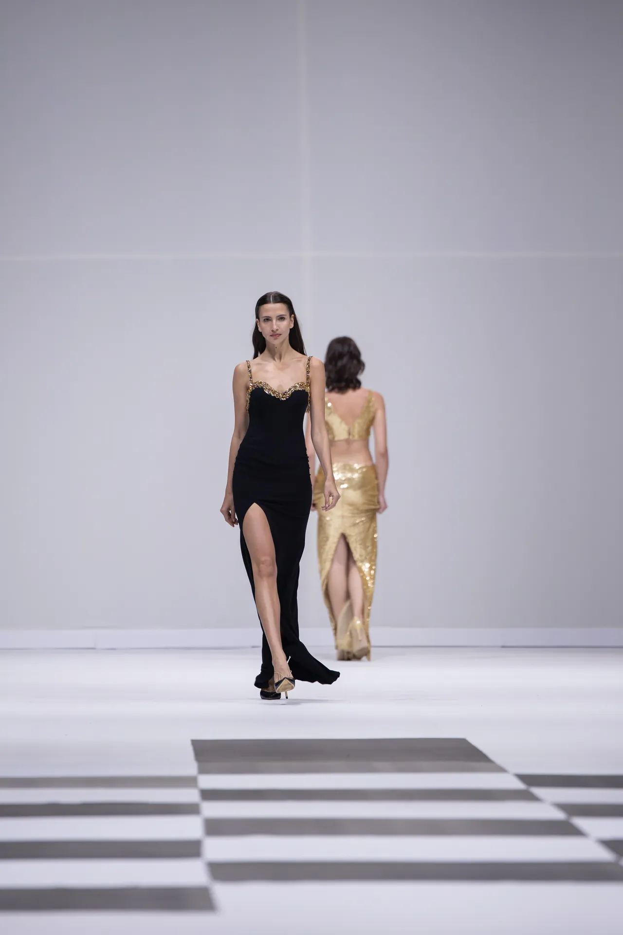 Model walks the runway in a black evening gown with gold trim and a high slit; another model in a gold sequined dress follows in the background.