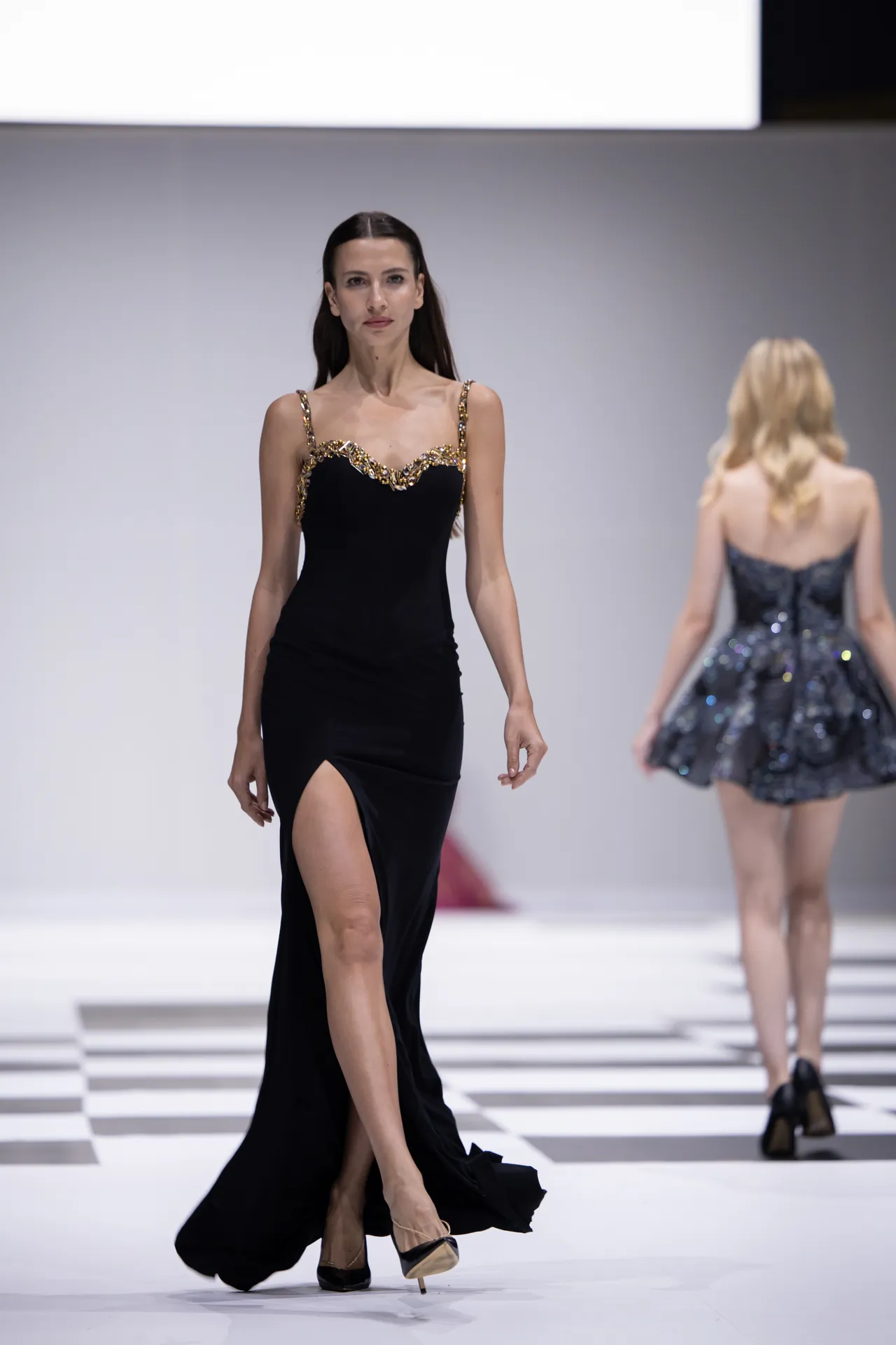 Model walks the runway in a black evening gown with gold-embellished straps and a high slit.