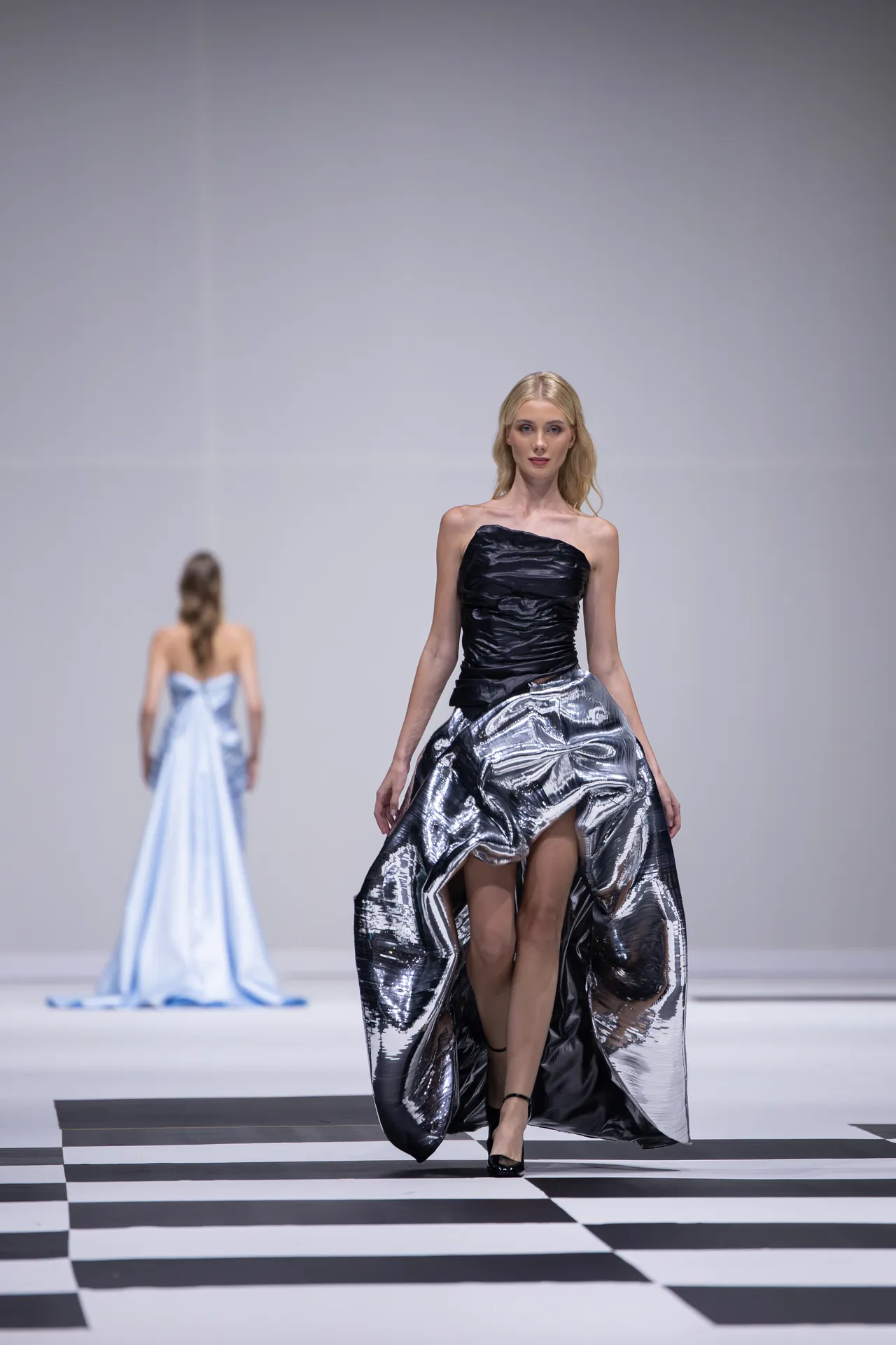 Model on a runway wearing a strapless black top with a shiny metallic silver skirt, walking forward on a black-and-white checkerboard floor; another model in a blue gown is blurred in the background.